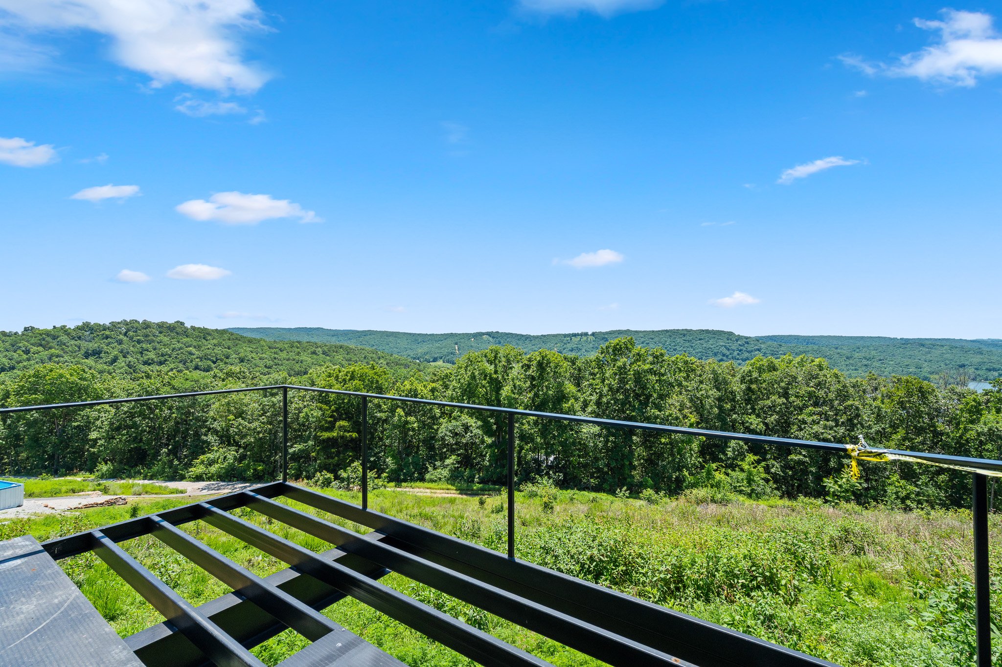 A scenic view of green forests, rolling hills, and a bright blue sky with some white clouds, seen from a balcony with black metal railing and partially assembled outdoor furniture.