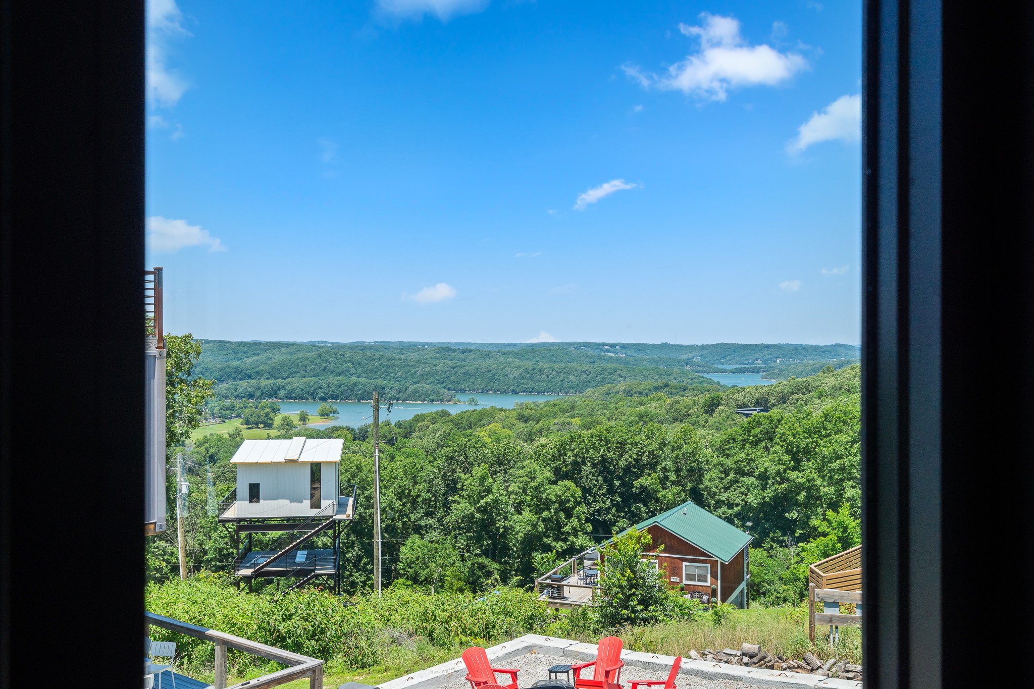 View of a river and green landscape through a window with dark frame. There are red chairs on a patio in the foreground, and houses with green roofs below, with trees and hills in the distance under a blue sky with a few clouds.