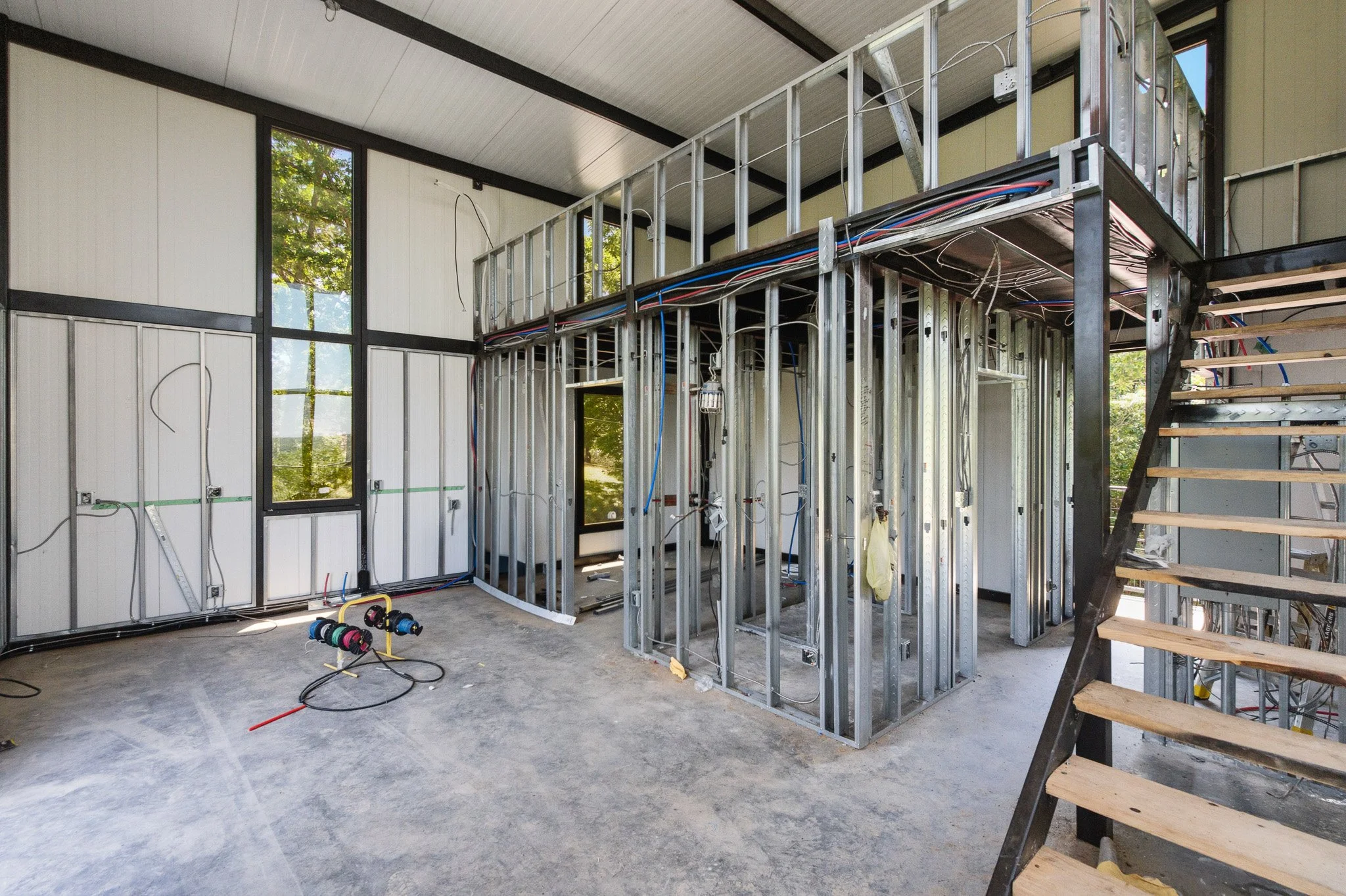 Interior of a house under construction, showing metal framing, electrical wires, and partially installed wall panels with large windows.