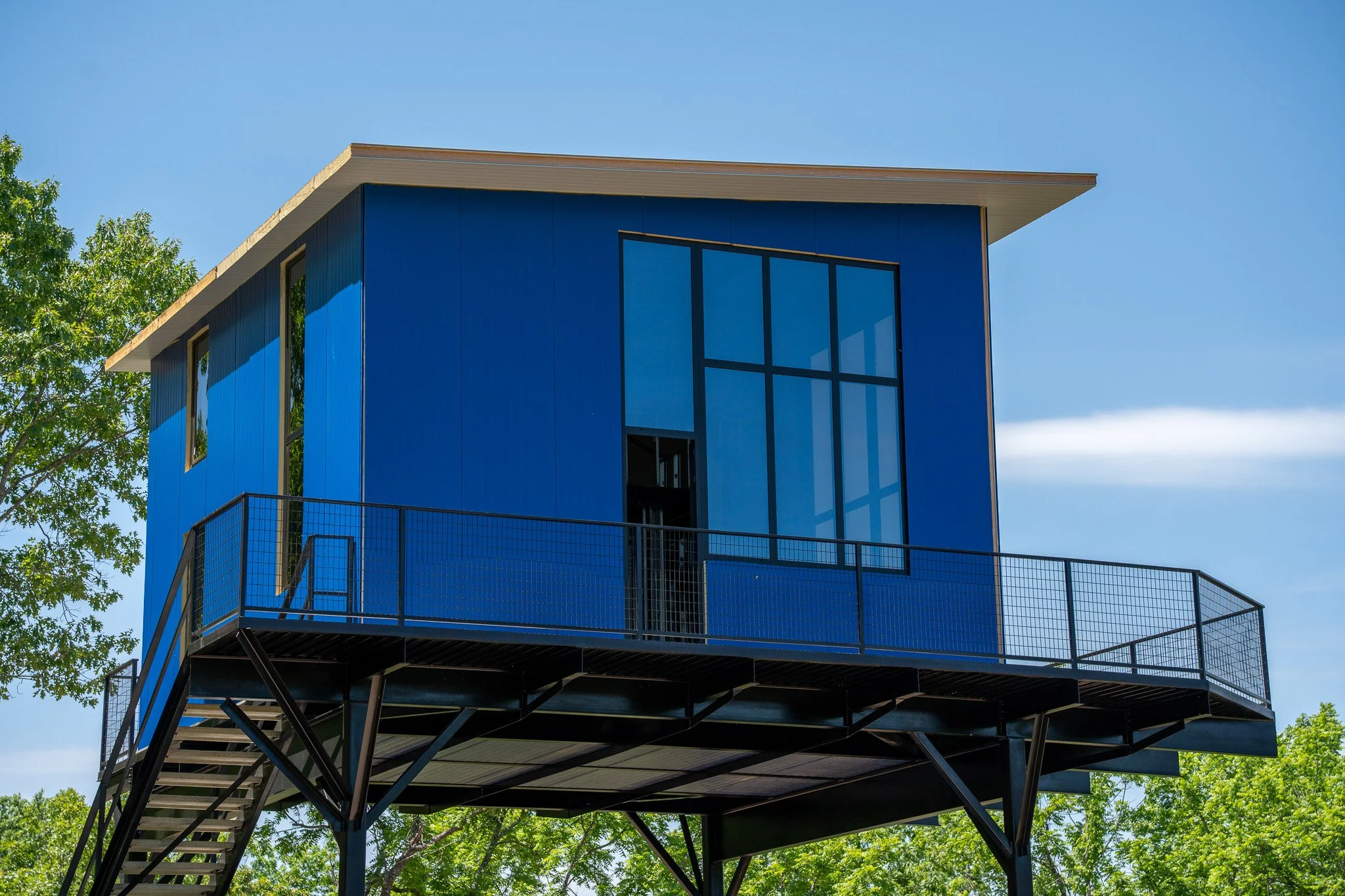 Blue modern house with large windows on a raised black metal structure, surrounded by green trees under a clear blue sky.