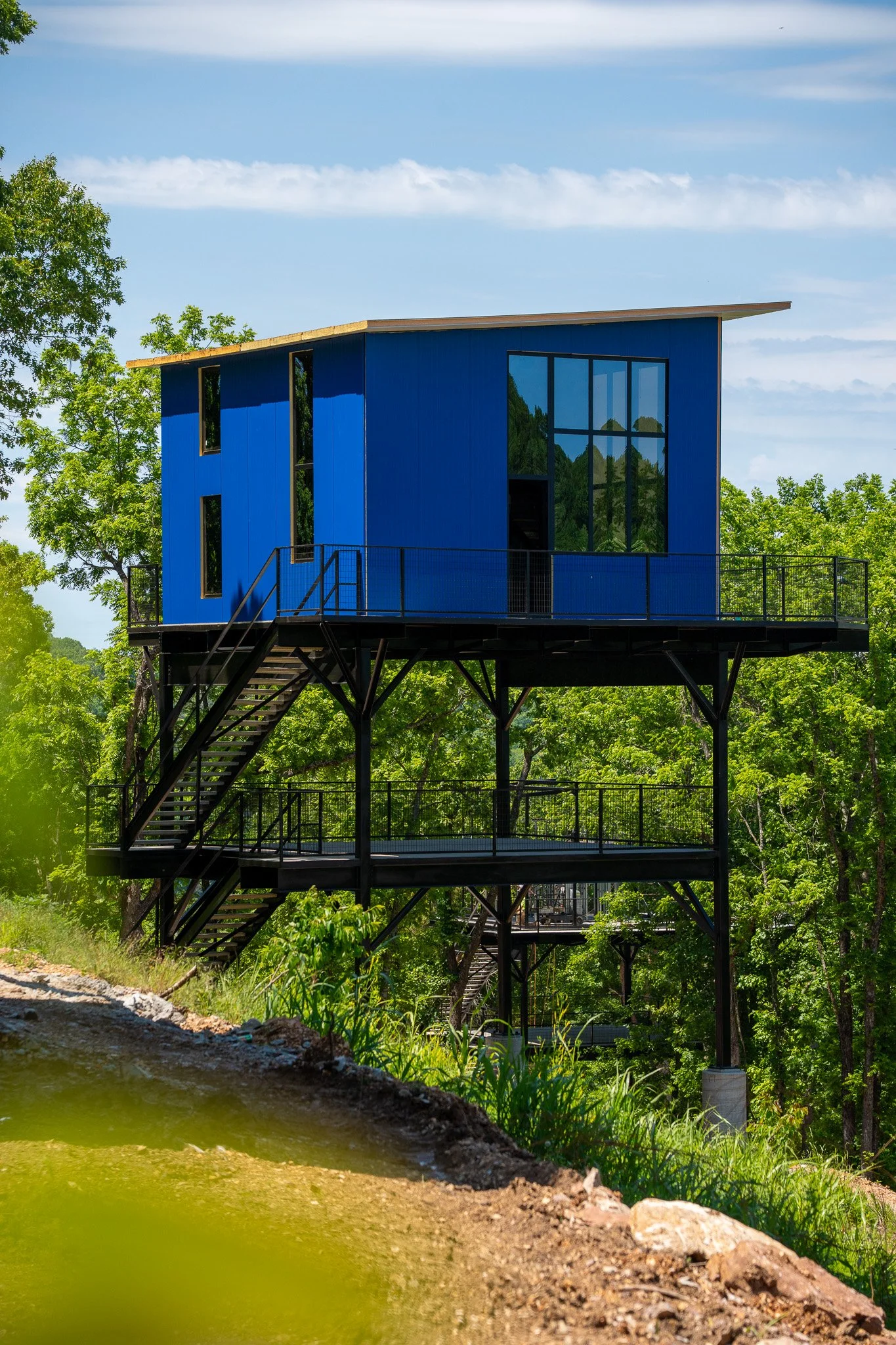 A modern blue house on stilts with large glass windows, surrounded by green trees under a partly cloudy sky.