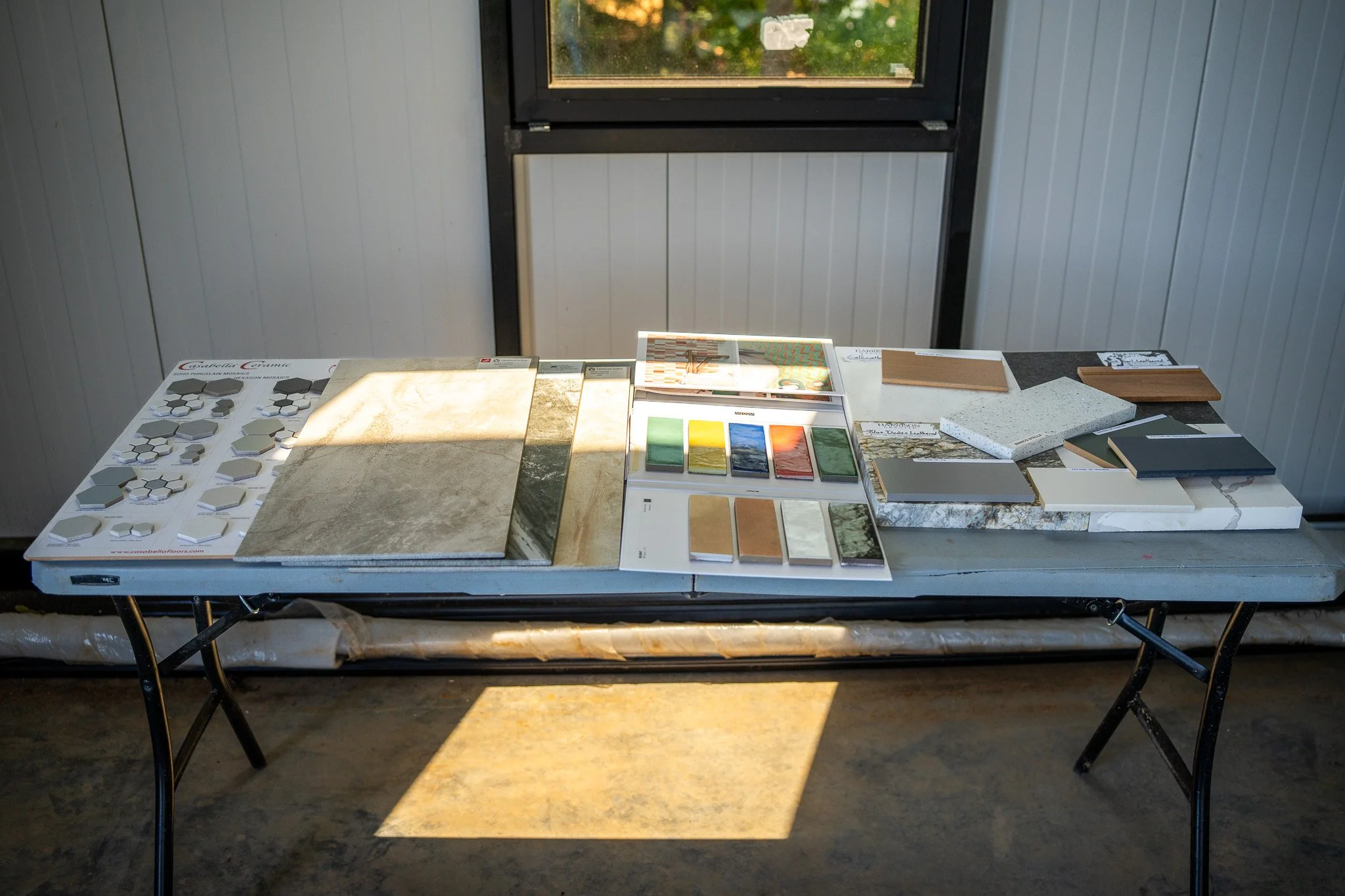Display of various tile samples and color swatches on a table inside a room, with a window in the background.