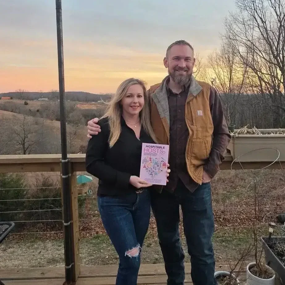 A woman and a man standing on a wooden deck outdoors at sunset, smiling, with the woman holding a book titled 'Hospitable Hosts.'