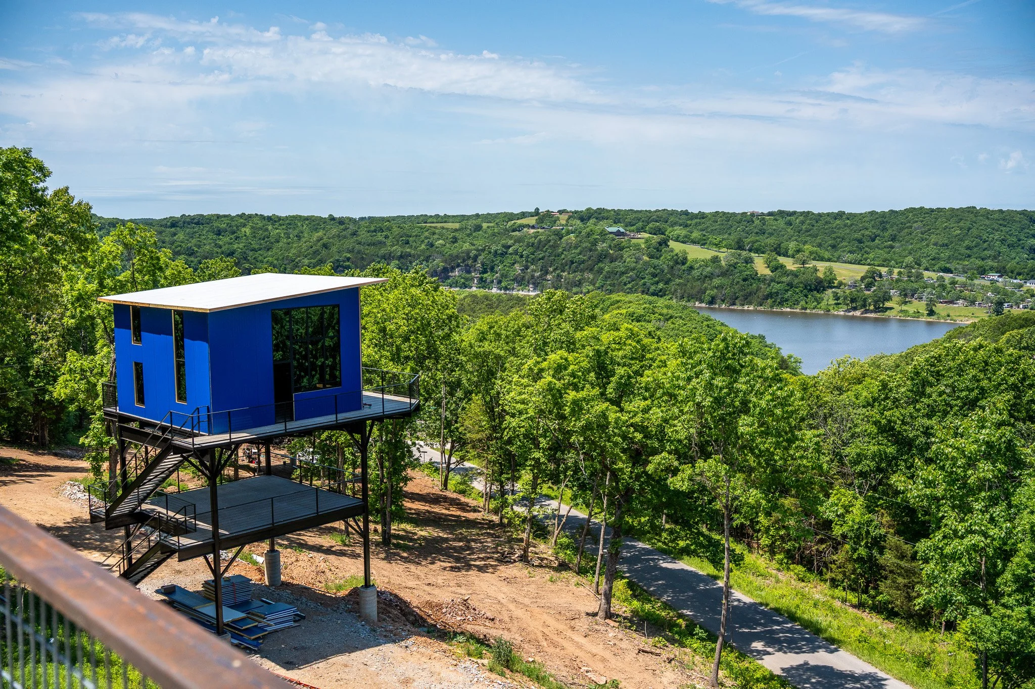 A scenic view of a green landscape featuring a river, rolling hills, and a blue sky with scattered clouds. In the foreground, there is a modern blue building on stilts with outdoor stairs, and trees surrounding the area.