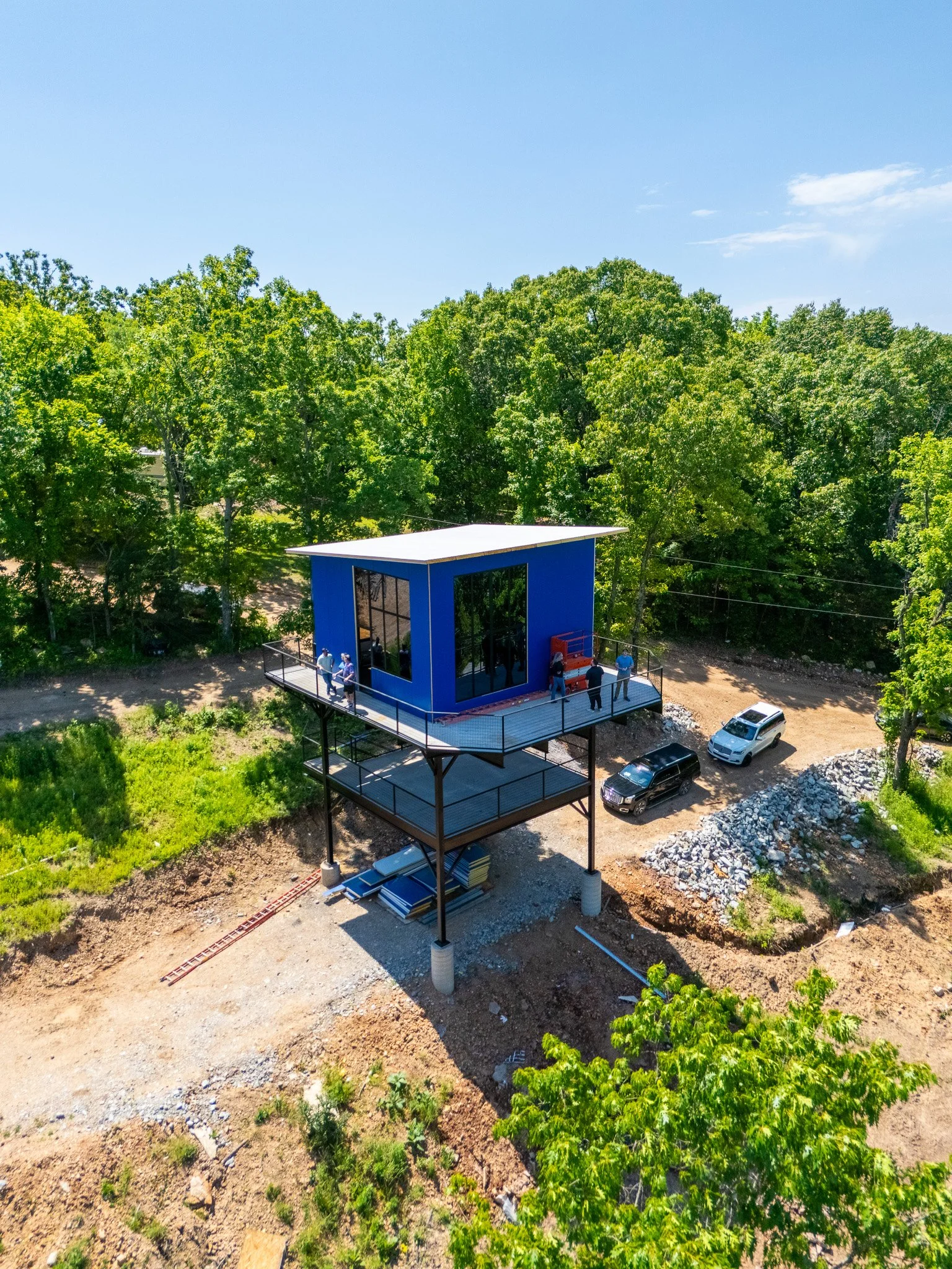 An elevated blue building with a flat roof and a large glass window, surrounded by green trees, parked cars, and construction site elements on a sunny day.