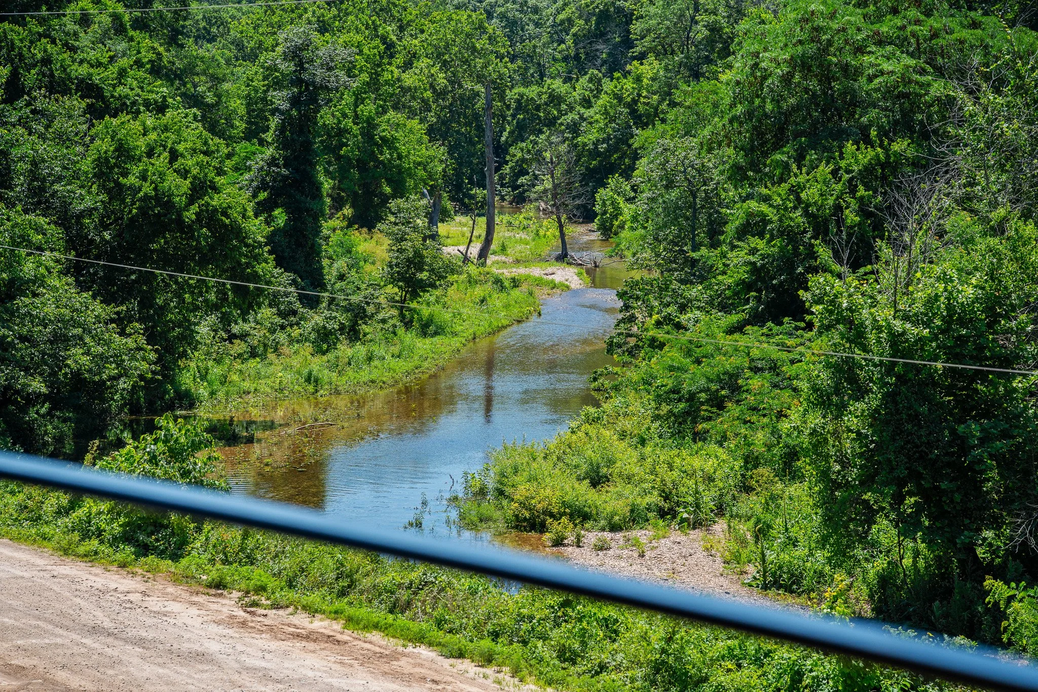 A winding river flowing through a lush green forest with trees and bushes, with a dirt road and a wire at the bottom of the image.