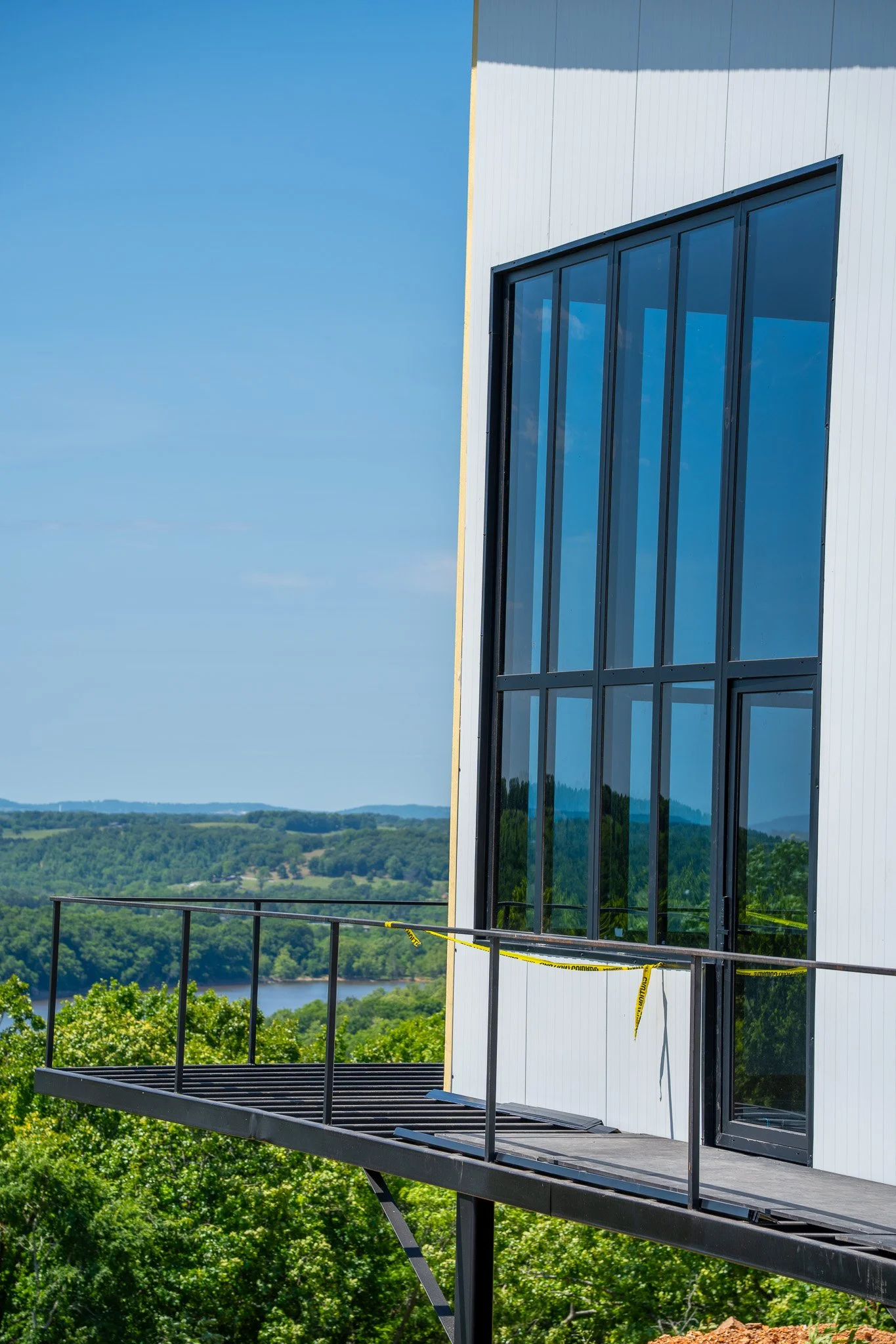 Modern building with large glass windows and a balcony overlooking a green landscape with trees and a river.