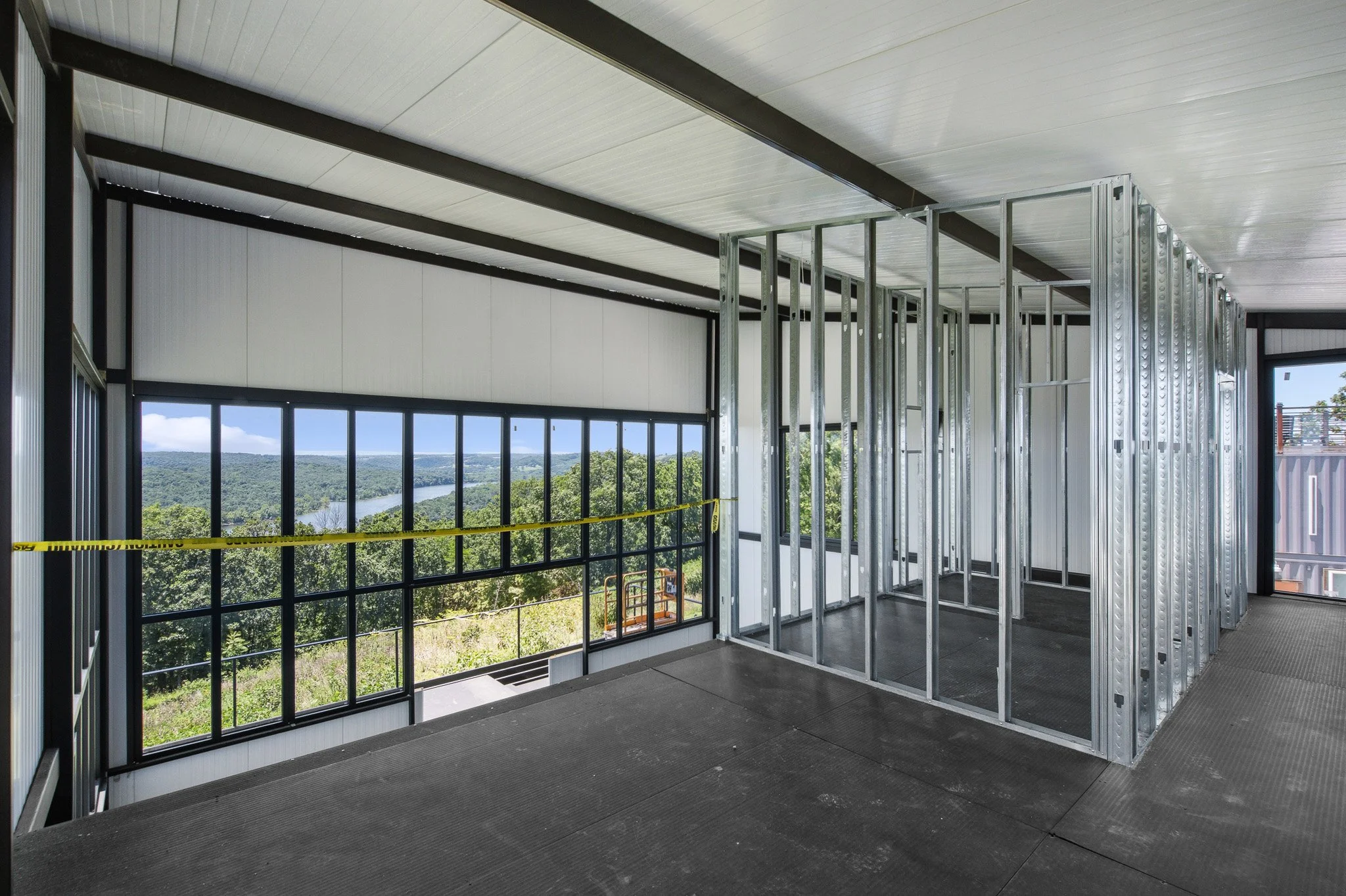 Interior of a building under construction with metal framing, large windows showing a river and trees, and a ceiling with white panels.