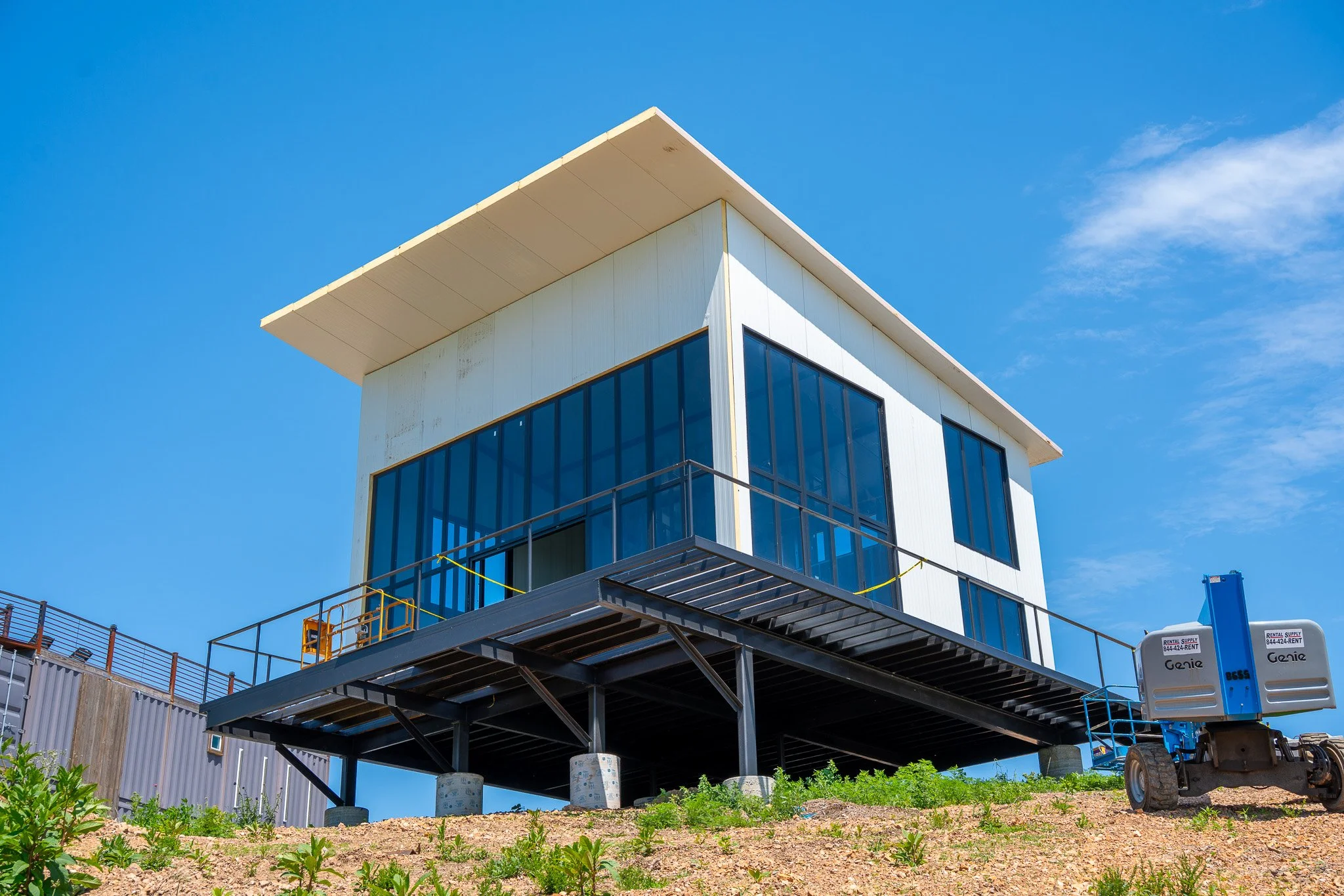 A modern building under construction with a distinctive flat overhanging roof, large glass windows, and construction equipment around the base, set against a bright blue sky with a few scattered clouds.