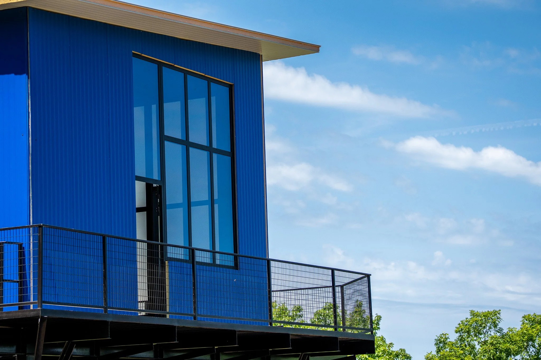 Modern blue building with large windows and a metal railing on the balcony, under a partly cloudy sky with some trees in the background.