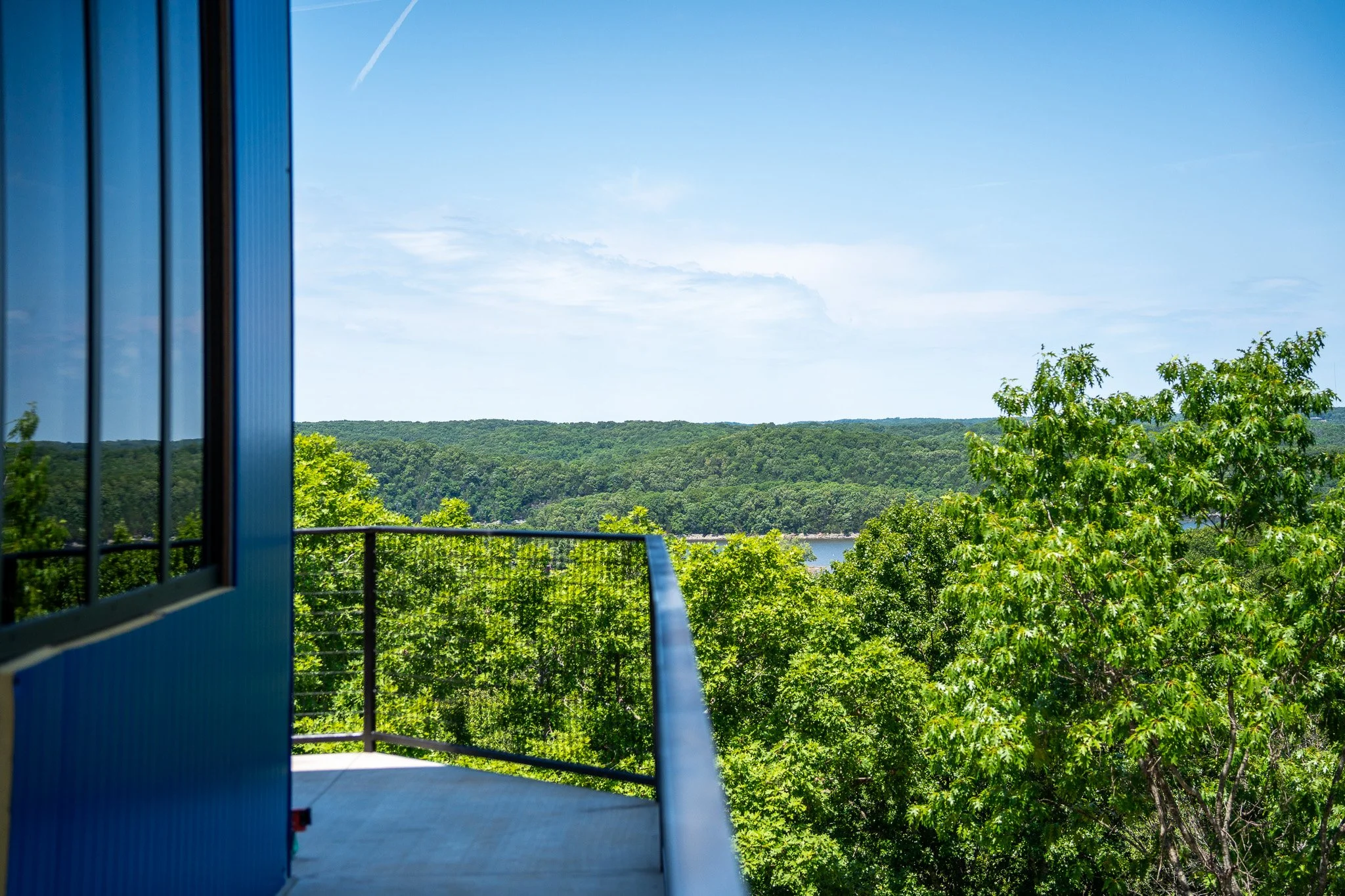 View from a balcony or deck overlooking a lush green forest and a river, with a blue building or structure partially visible on the left.
