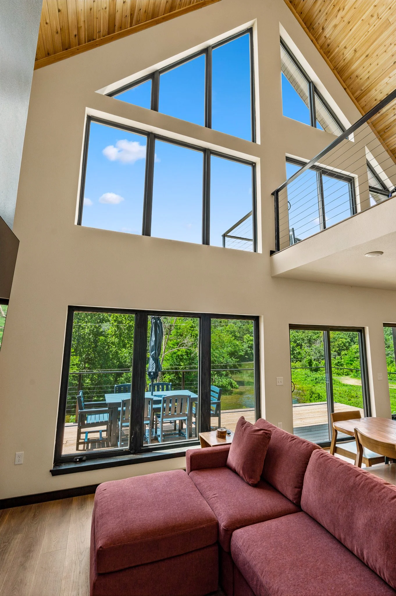 Interior of a living room with large windows showing an outdoor patio with a table and chairs, and a view of trees and a river.