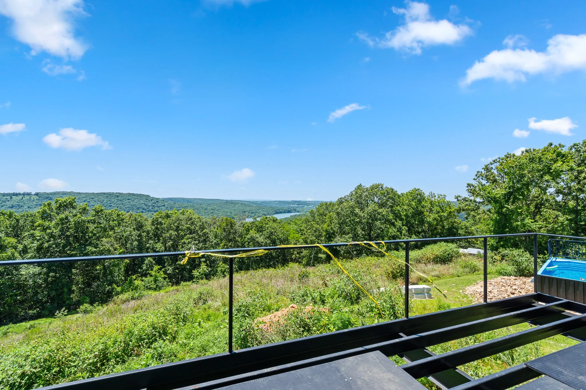 A scenic view of a lush green landscape with trees and hills under a bright blue sky with scattered white clouds, seen from a balcony with a black metal railing and caution tape.