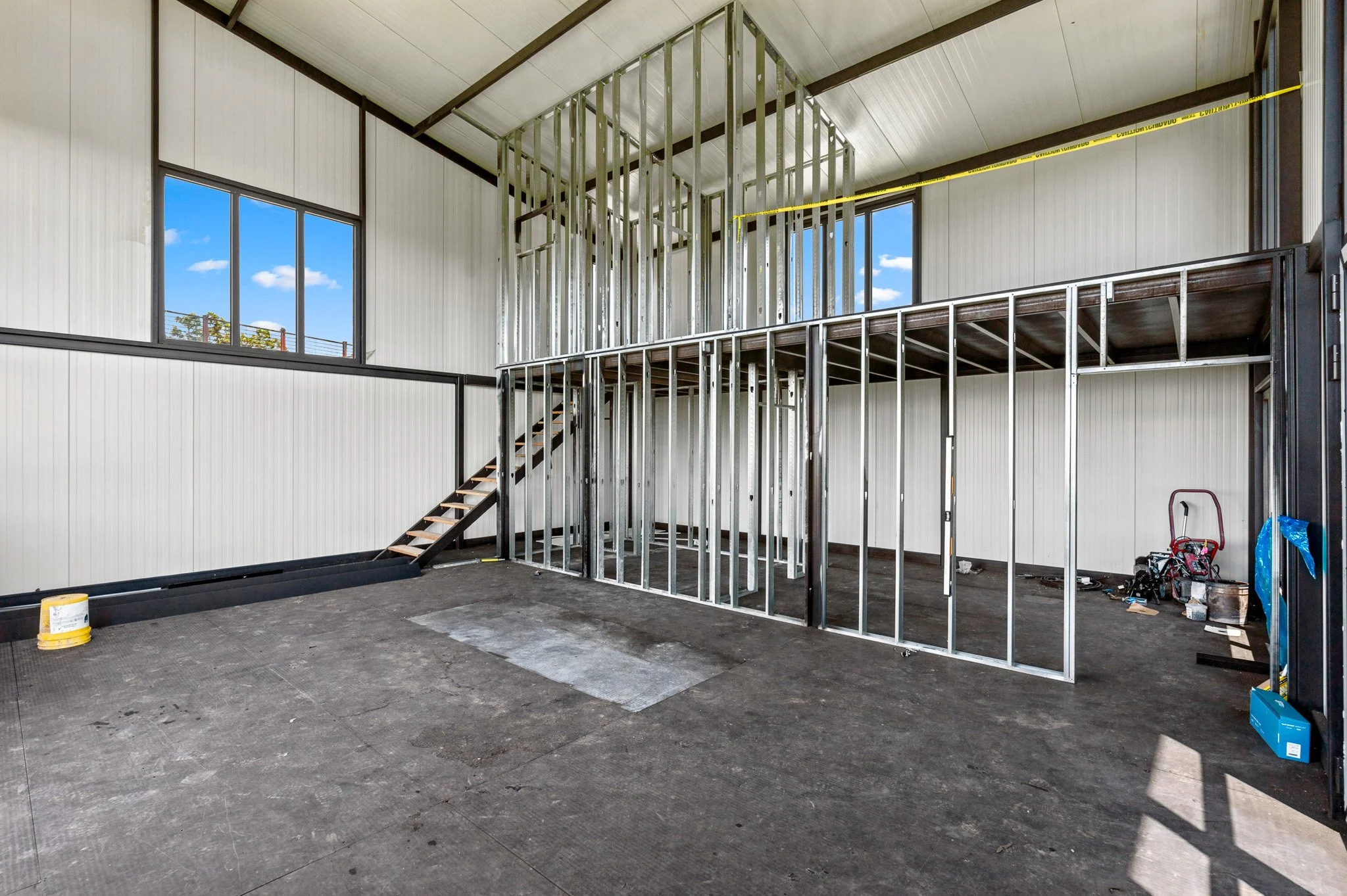 Interior of a building under construction with metal framing for walls and a staircase leading to a second floor, with large windows and construction tools on the floor.
