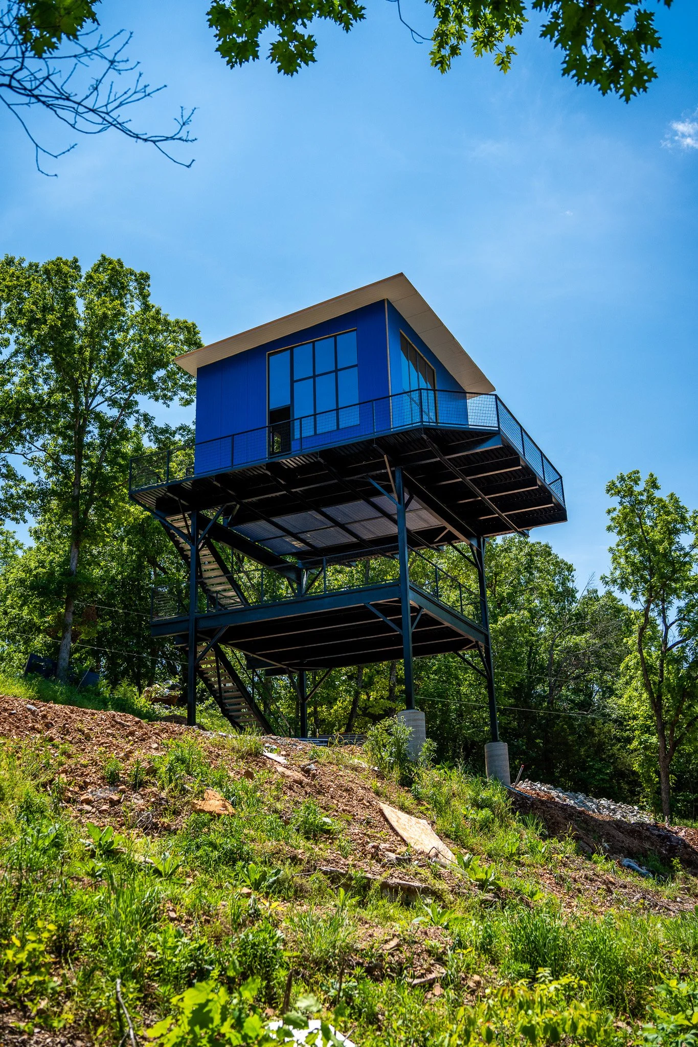 A modern tiny house painted blue, elevated on metal stilts with outdoor stairs and a balcony, surrounded by trees and greenery under a clear blue sky.
