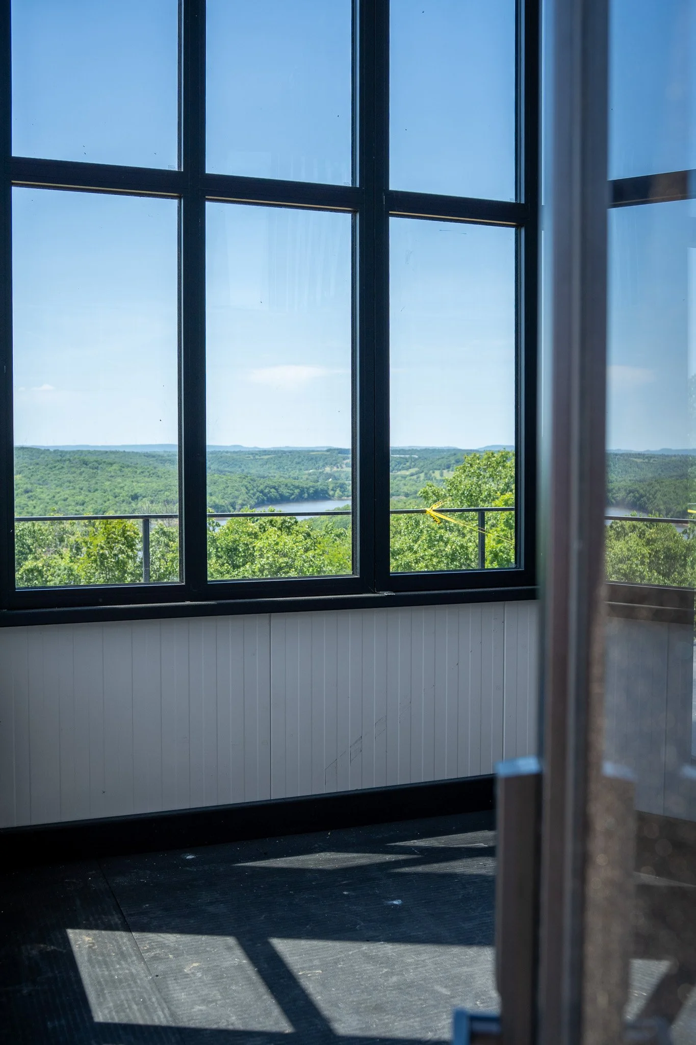 View of a landscape through large windowpanes with black frames, showing green trees and distant hills under a clear blue sky.