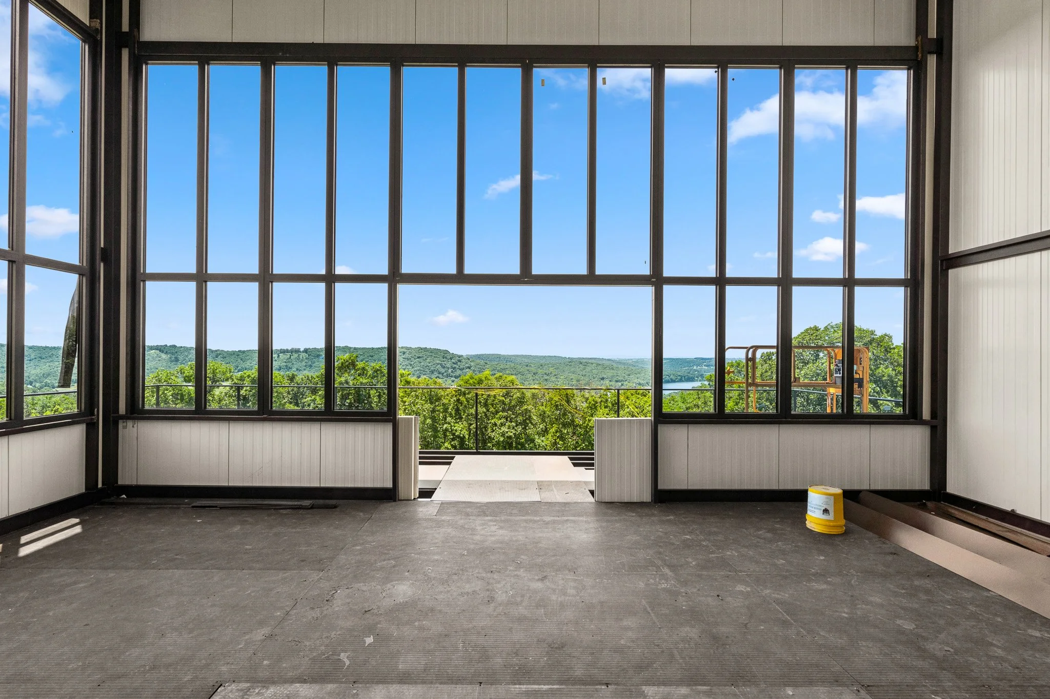 Empty room under construction with large window showcasing a scenic view of green trees and a river, construction tools and materials present.
