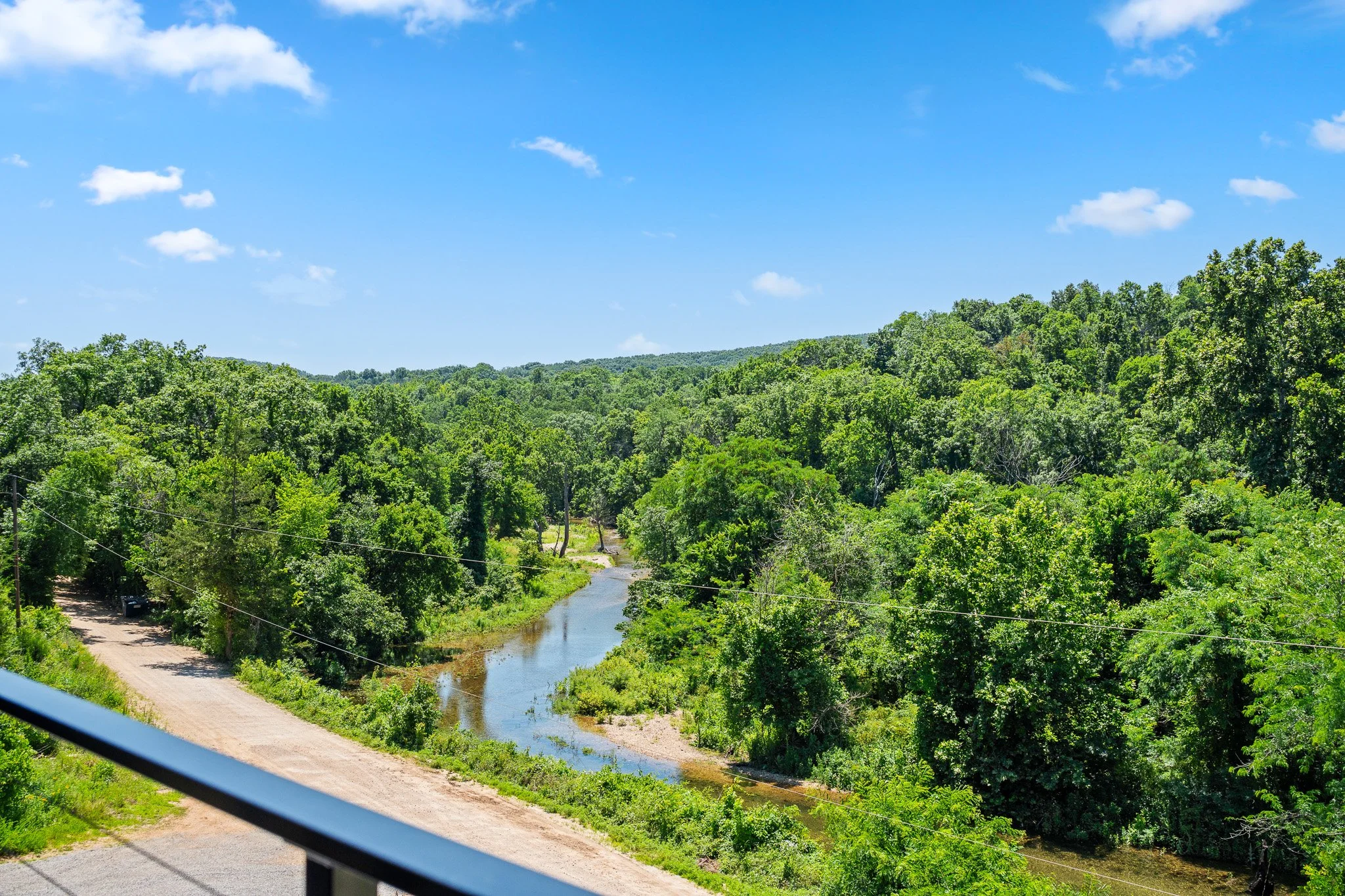 View of a lush green forest with a small river running through it, seen from a vehicle window on a bright, partly cloudy day.