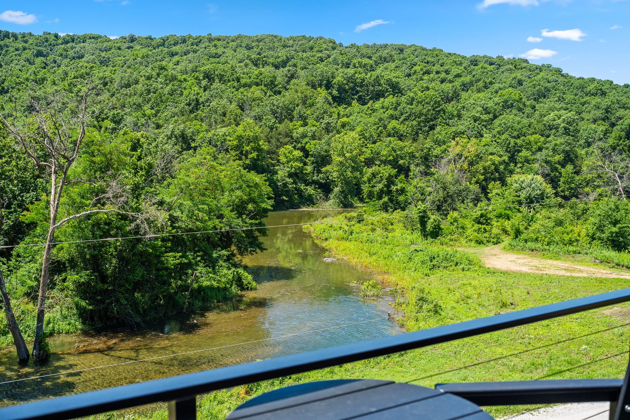 A scenic view of a lush green forested hillside reflecting in a river, with a small dirt path and a blue railing in the foreground on a sunny day.