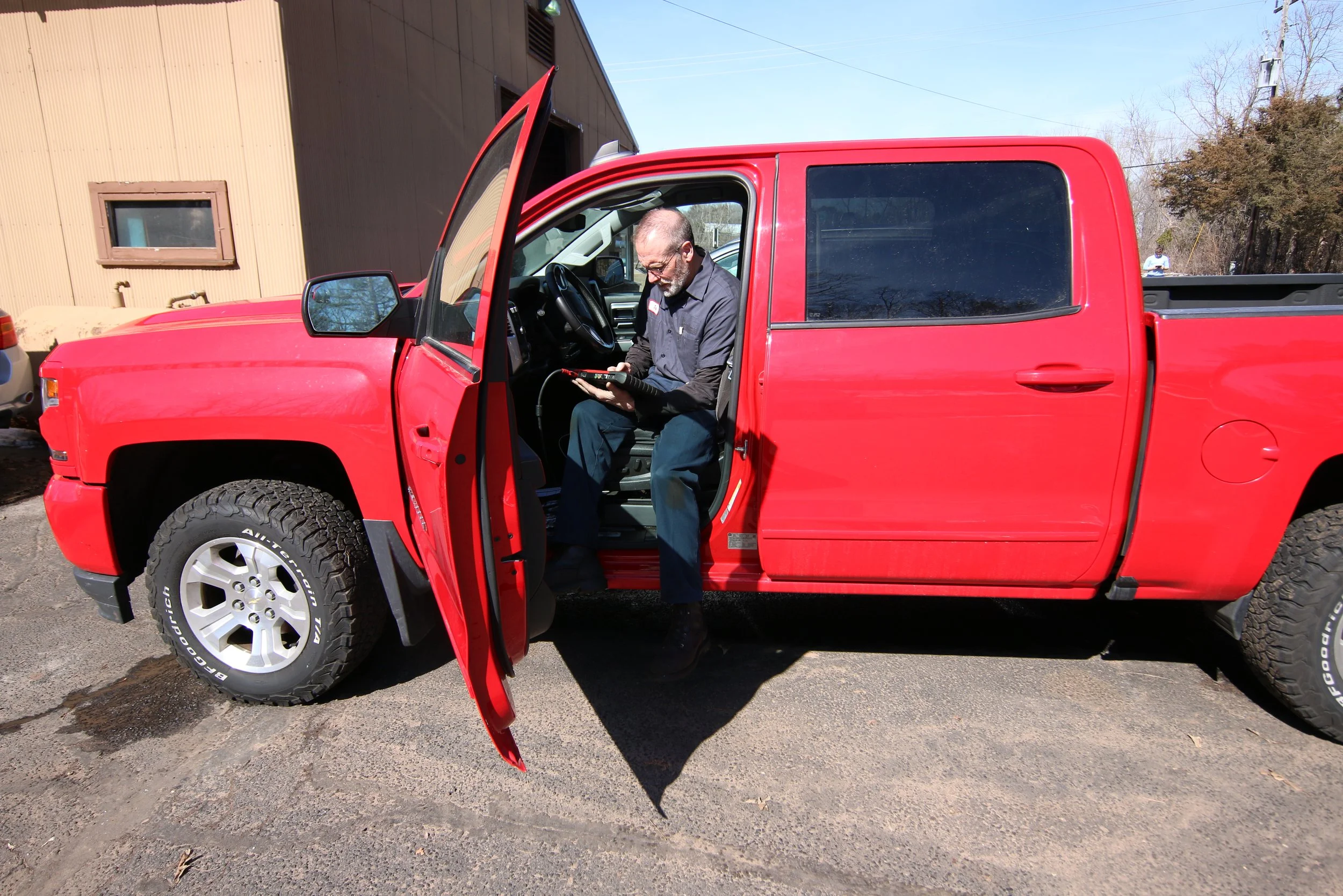 A man sitting in the driver's seat of a red pickup truck, looking at a handheld device, with the driver’s side door open.