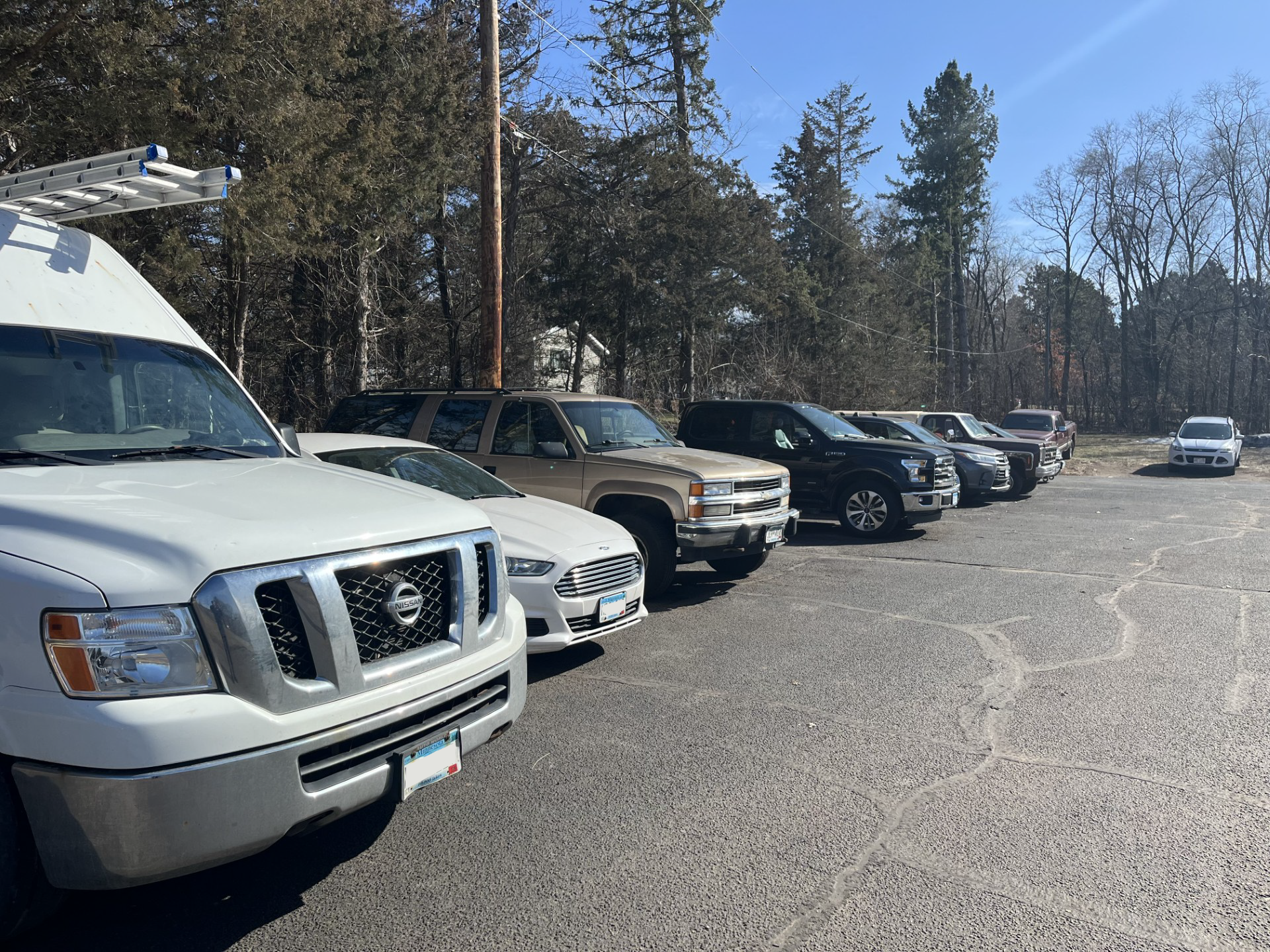 A row of parked vehicles including a white van, a white car, a brown truck, a black truck, a silver truck, a gray truck, and a small white car on a cracked asphalt lot with trees and clear blue sky in the background.