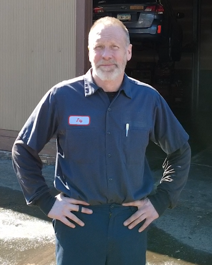 A man with a beard and short white hair standing outdoors in front of a garage, wearing a navy blue work uniform with a name tag that says 'Jay' and a pen in his shirt pocket, smiling with hands on hips.