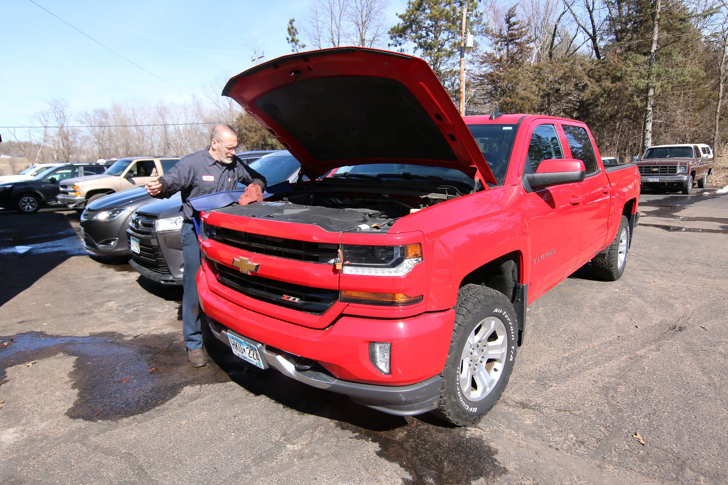 A man in mechanic overalls inspecting the engine of a red Chevrolet Silverado pickup truck with the hood open in a parking lot.