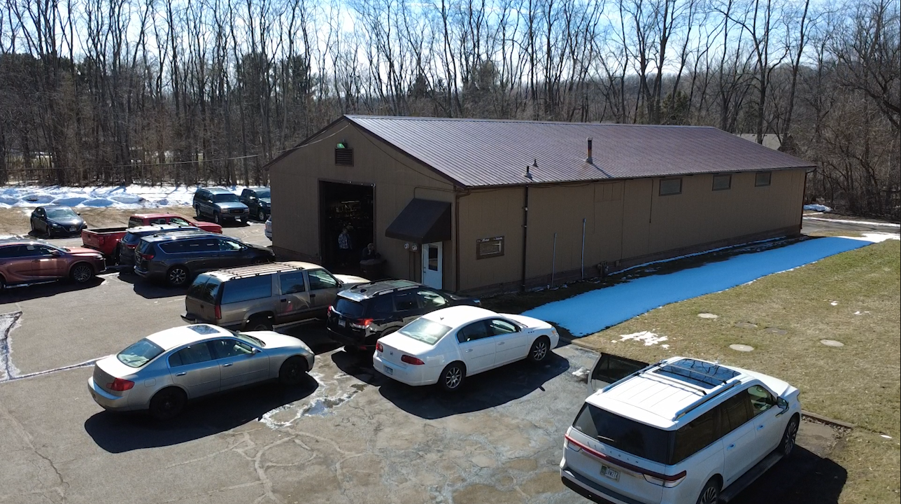 Parking lot with several cars parked outside a large beige building with a metal roof, surrounded by leafless trees and patches of snow on the ground.