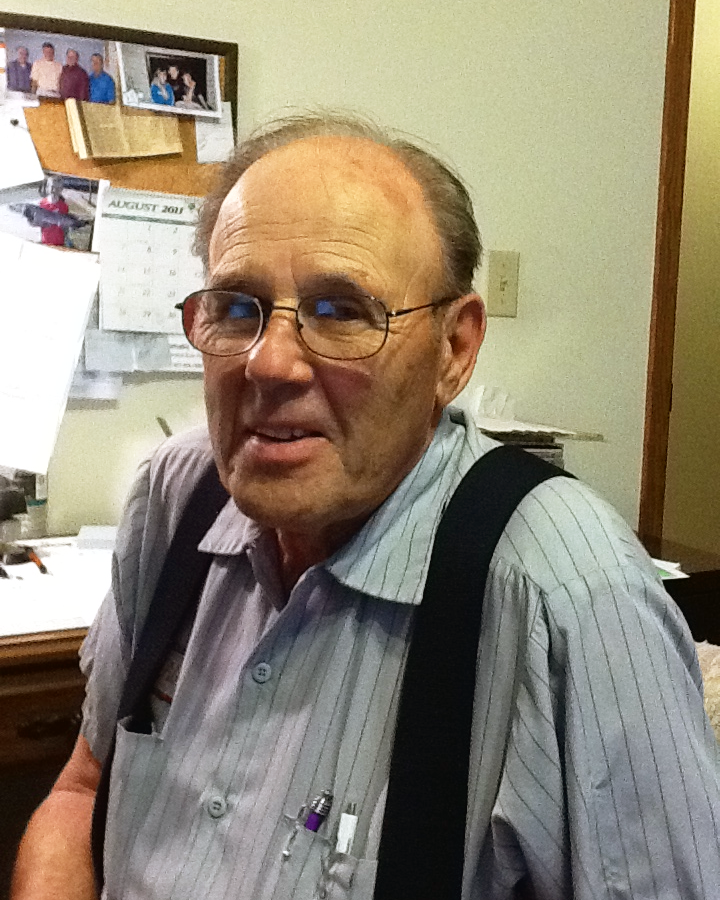 An elderly man with glasses and a striped shirt is smiling in an office, with a bulletin board and framed photos in the background.
