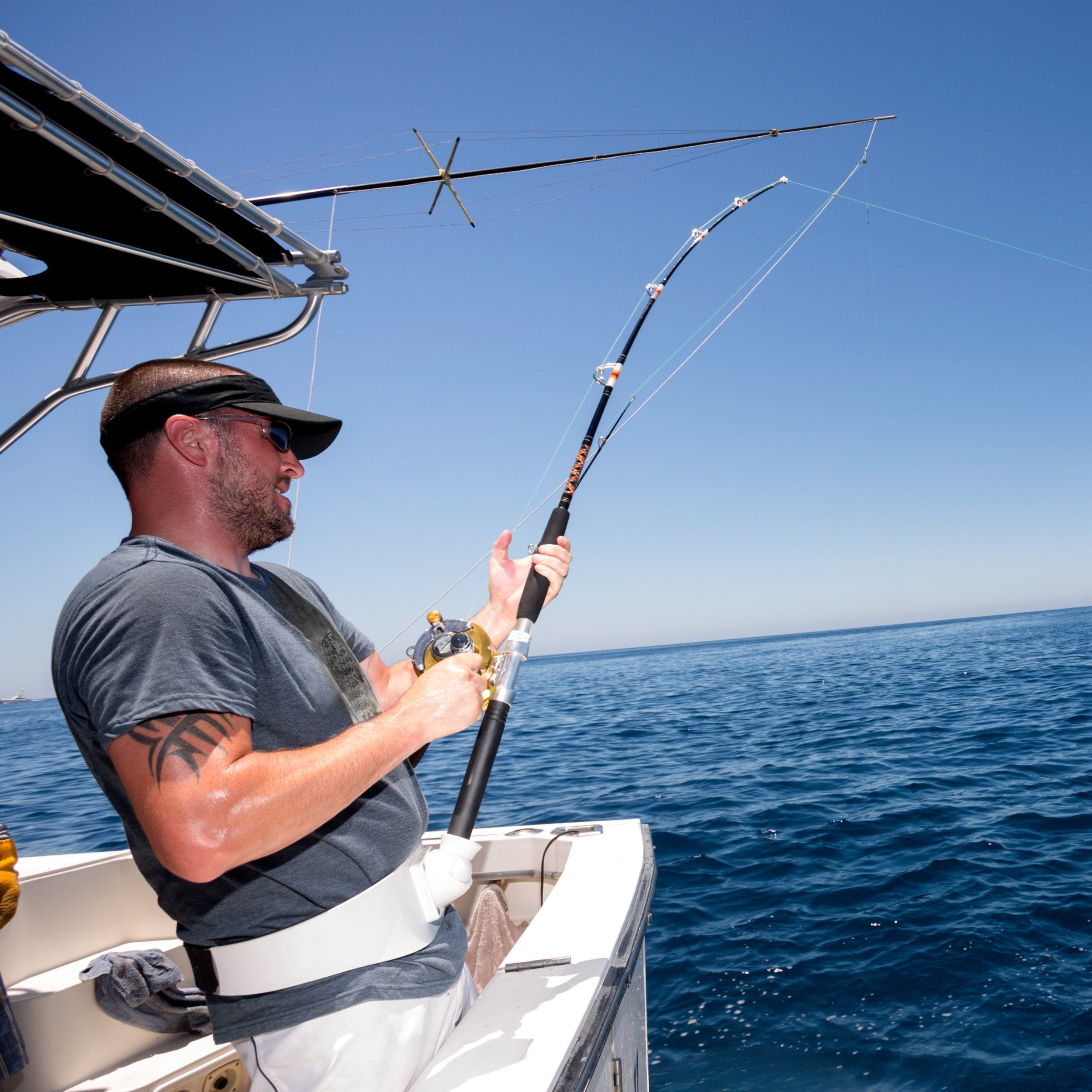 Man wearing sunglasses and a hat fishing on a boat in the ocean under a clear blue sky.