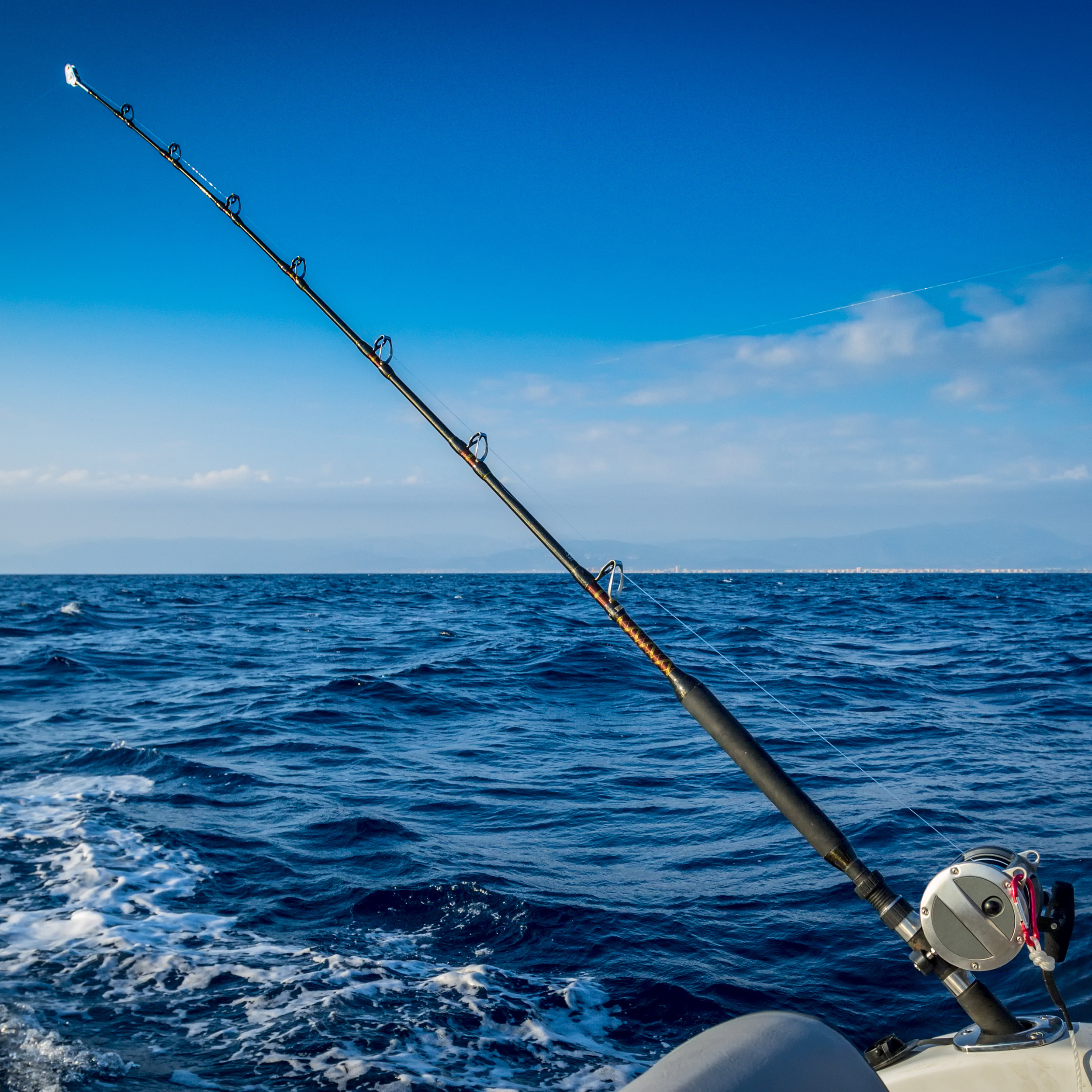 Fishing rod extending from a boat into the ocean during daytime with a distant shoreline under a blue sky.