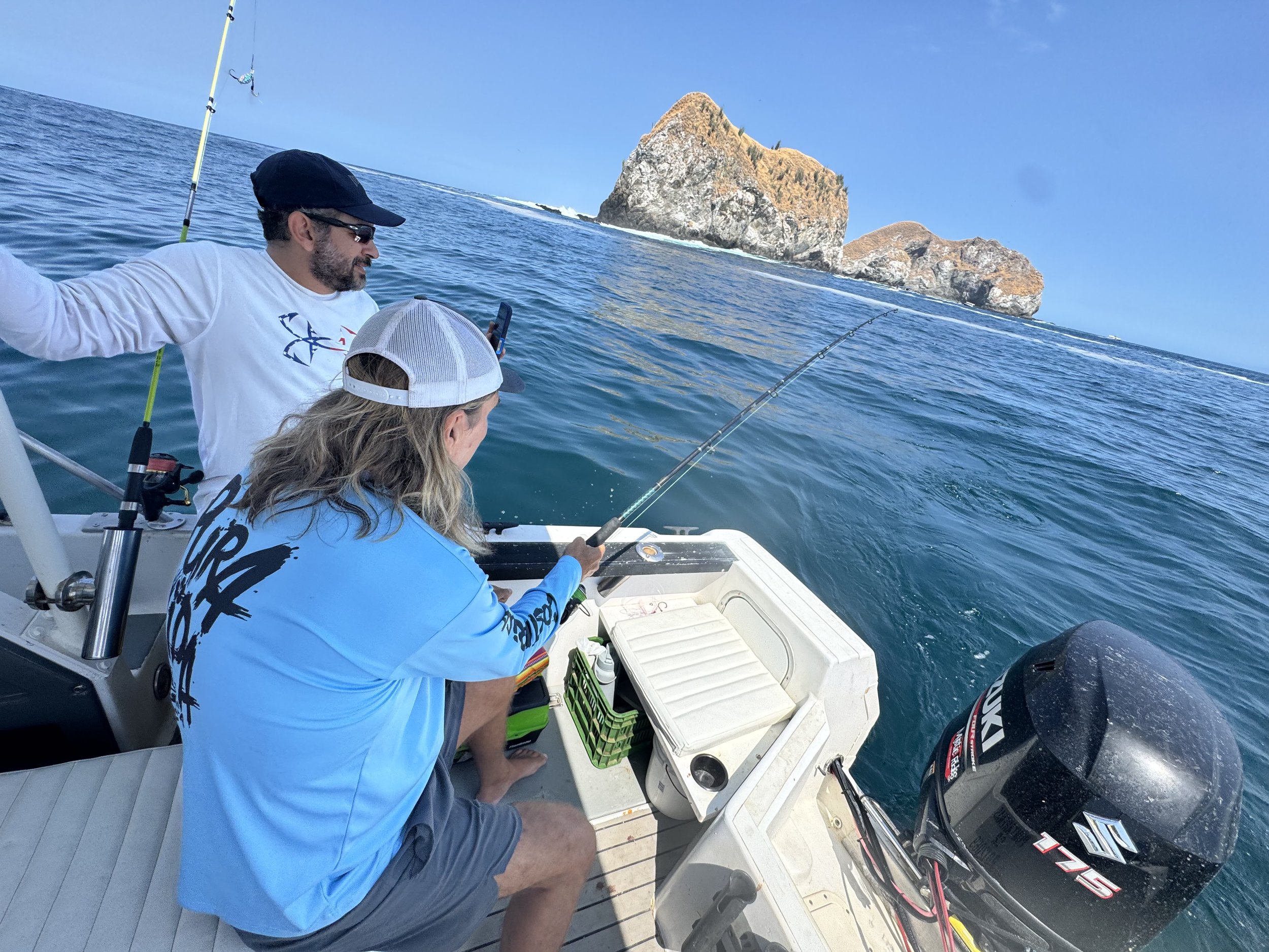 Two men fishing on a boat with large rocky islands in the background under a clear blue sky.