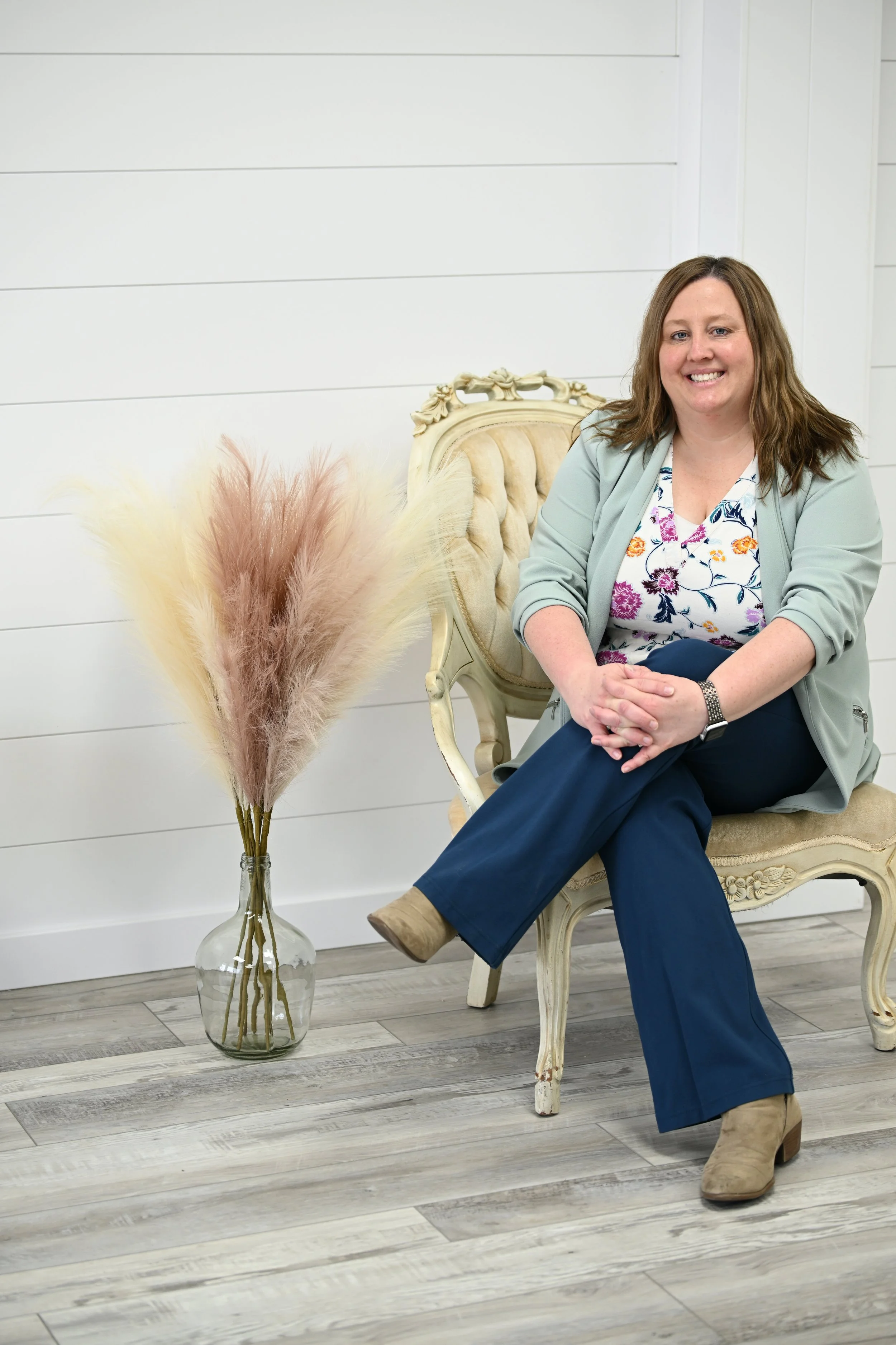 A woman sitting in a vintage-style armchair on a wooden floor, smiling, with a large vase of pampas grass next to her.