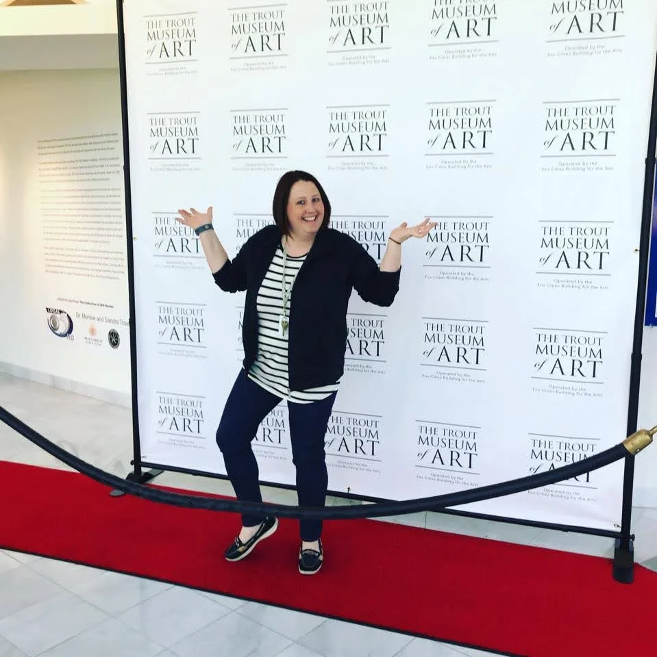 Woman standing on a red carpet in front of a backdrop with the text 'The Trout Museum of Art', smiling and posing with arms raised.