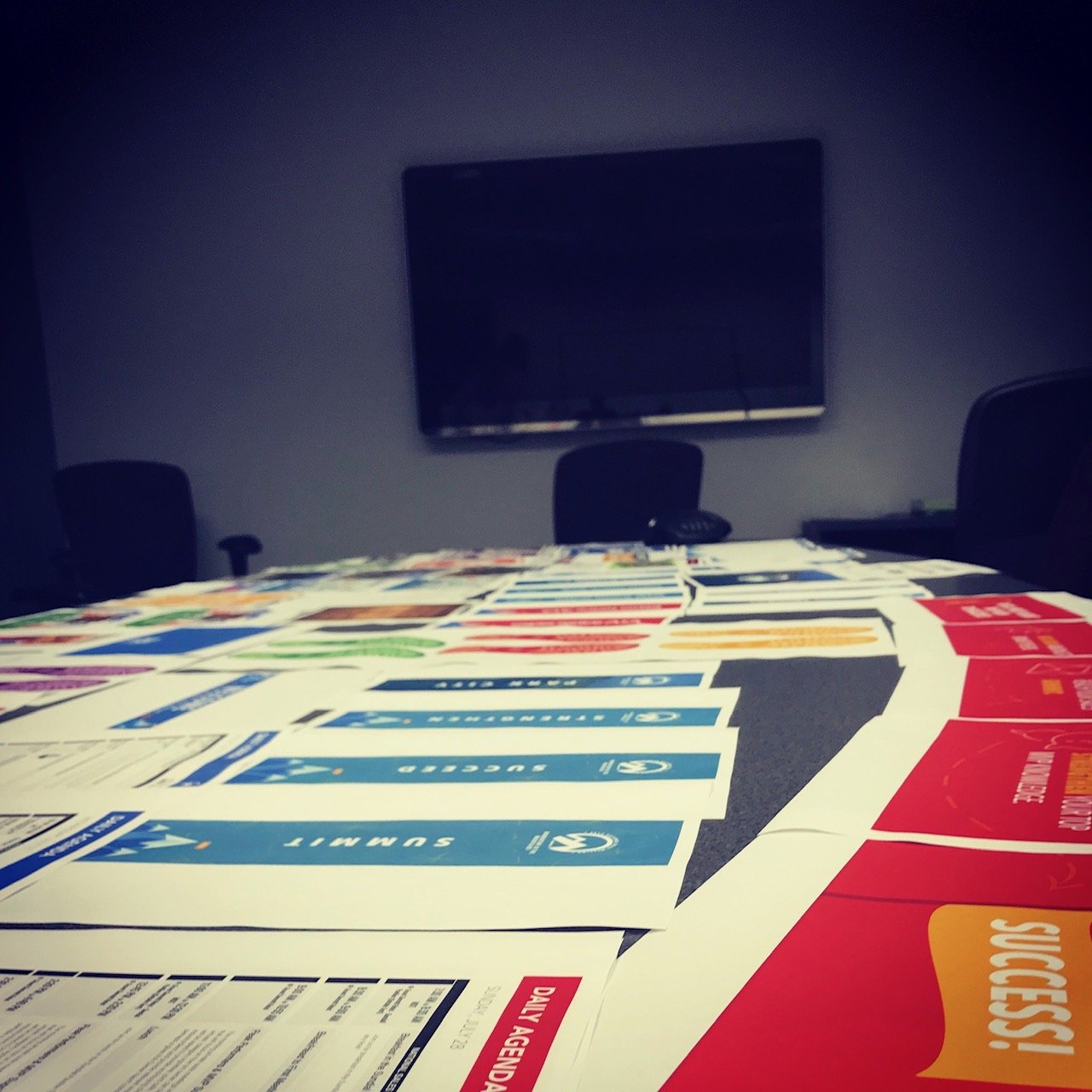 Table covered with colorful blue, red, green, and yellow paper materials, possibly flyers or brochures, in a conference room with a large TV on the wall and black office chairs around the table.