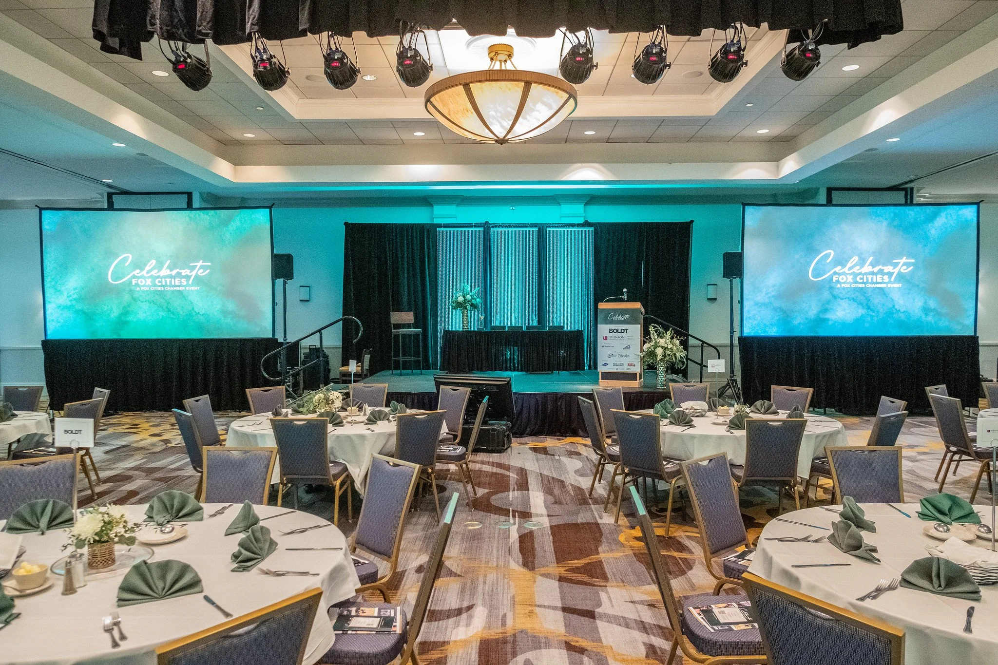 Conference room with round tables set for a meal, each table with green napkins folded in a fan shape, silverware, and floral centerpieces. A stage with two large screens displays the message 'Celebrate Fox Cities' and has black curtains, flowers, and a podium.