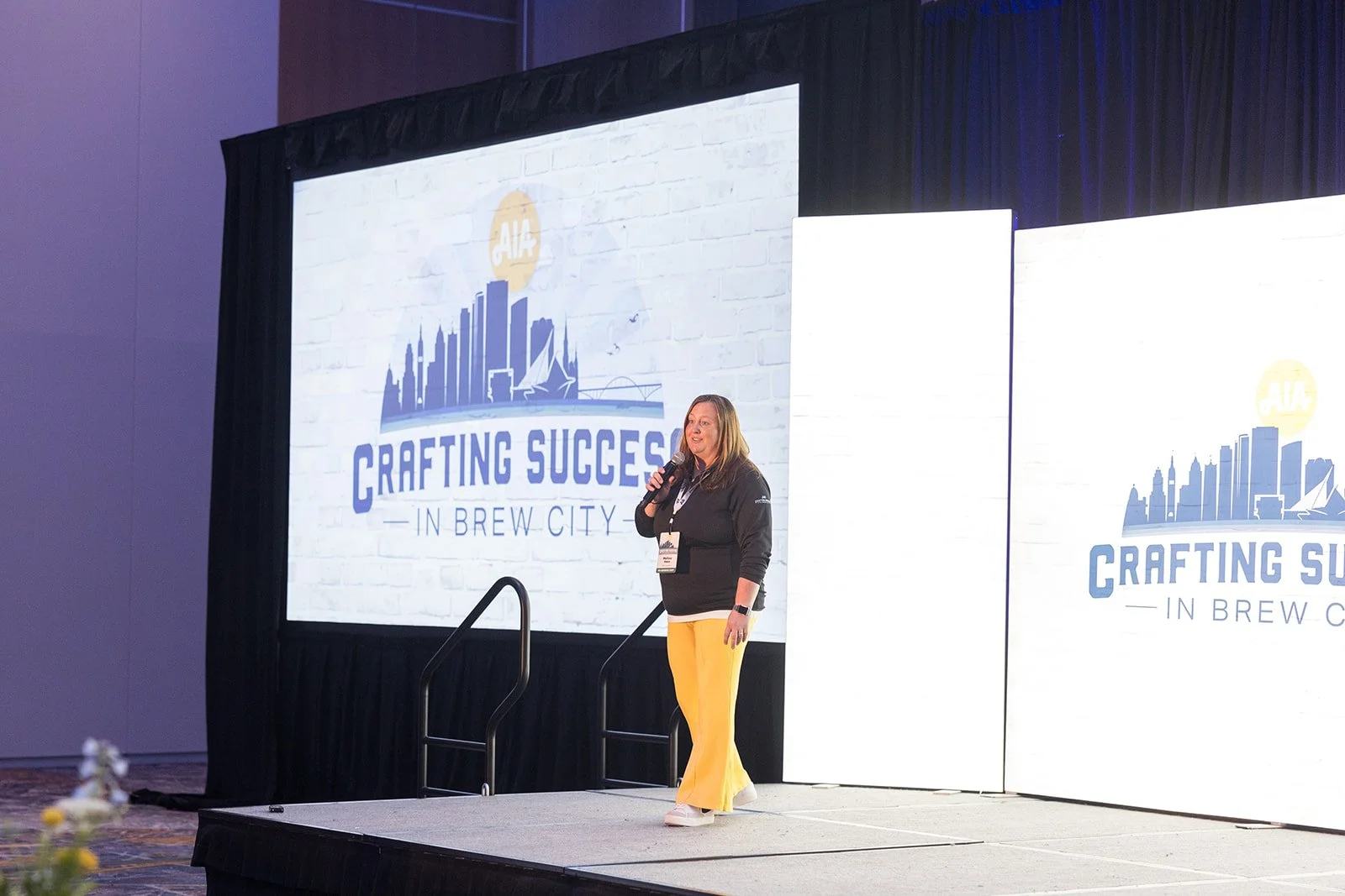 A woman standing on stage speaking into a microphone at a conference, with a large screen behind her displaying a logo for 'Crafting Success in Brew City' and an illustrated skyline.