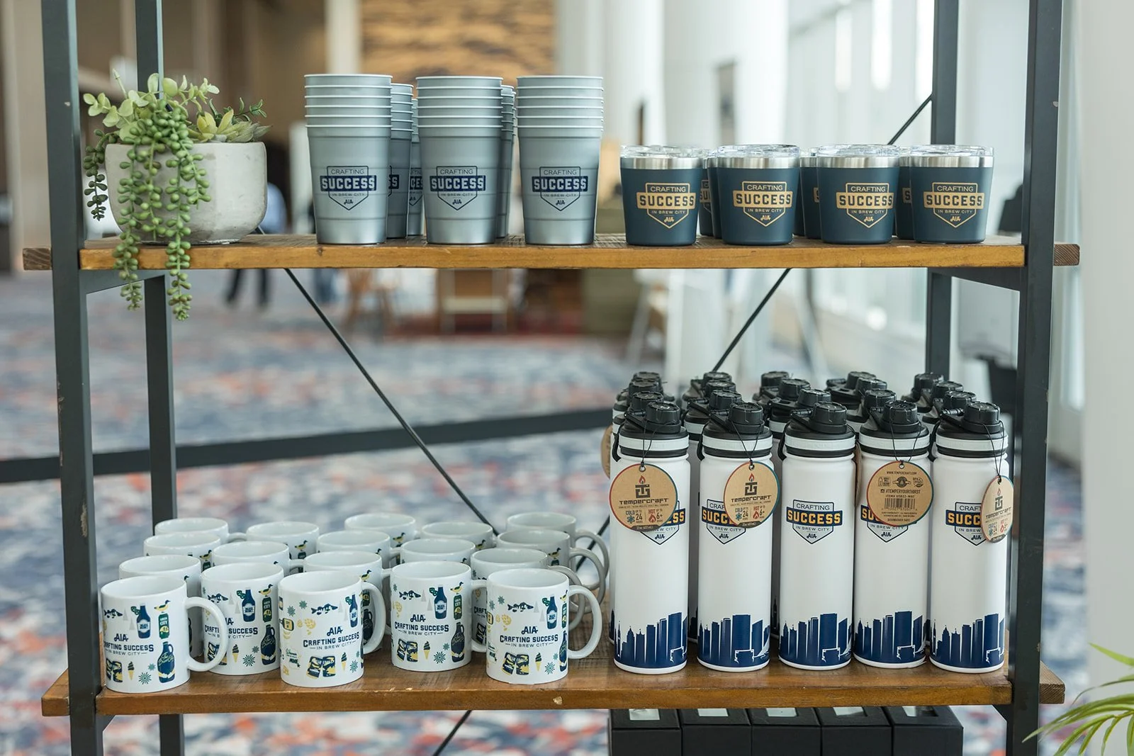 A display shelf with reusable cups, mugs, and water bottles featuring branding for a craft brewing event called Crafting Success in Brew City.