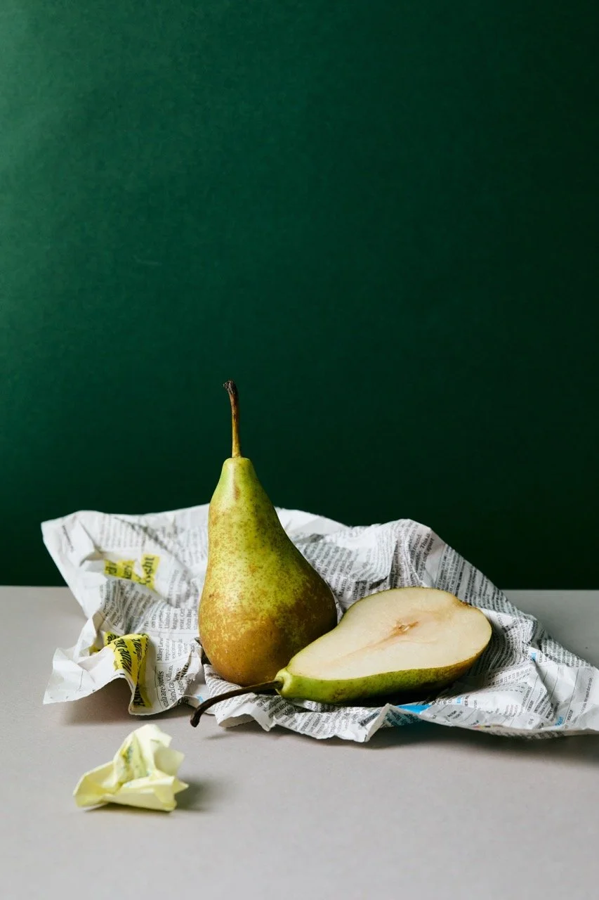 A whole pear and a cut pear on a crumpled newspaper, with a small crumpled piece of paper in front, against a green background.