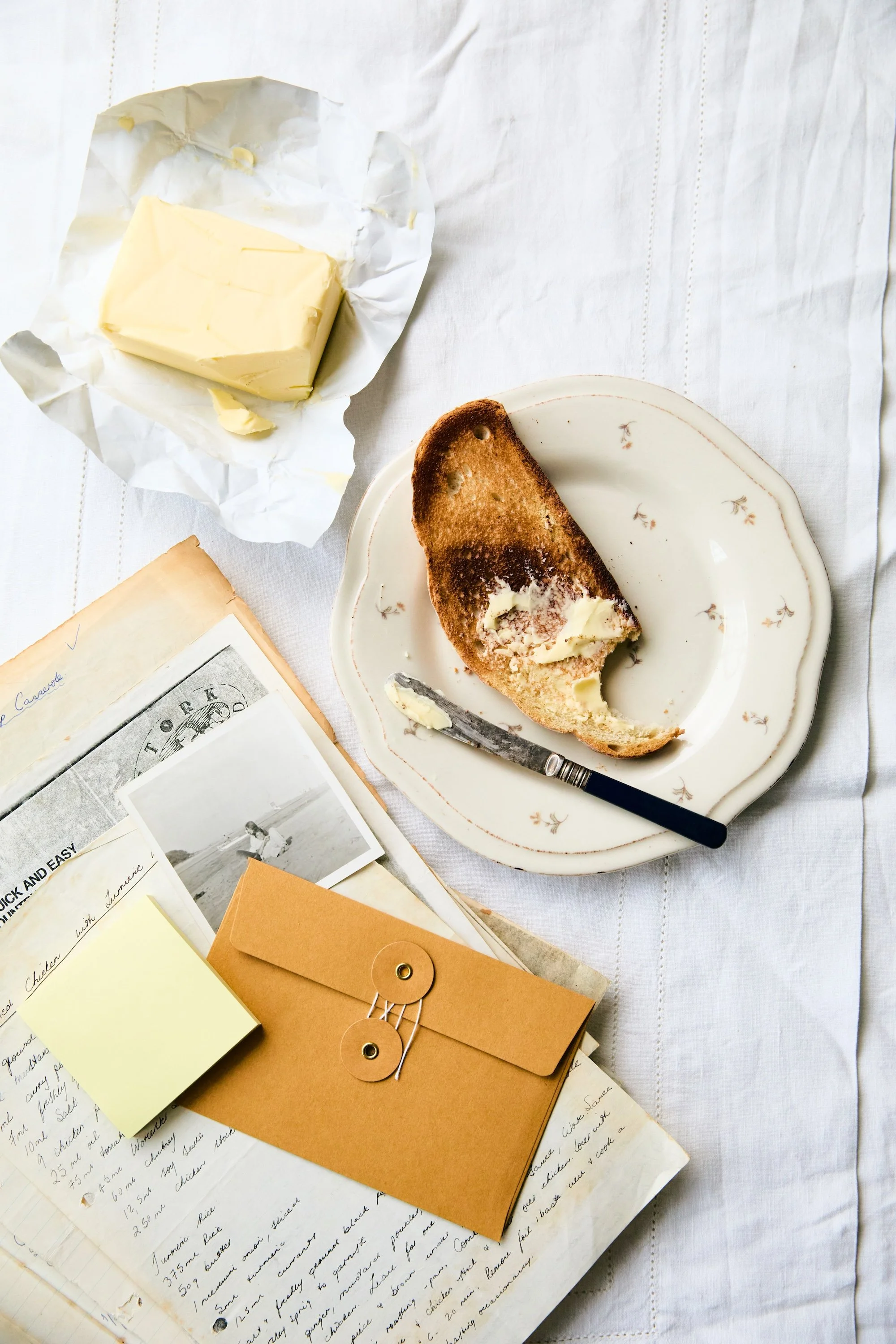 An overhead shot of a table with half eaten toast, a block of butter and an open book with handwritten recipes, a photograph and a manilla envelope.