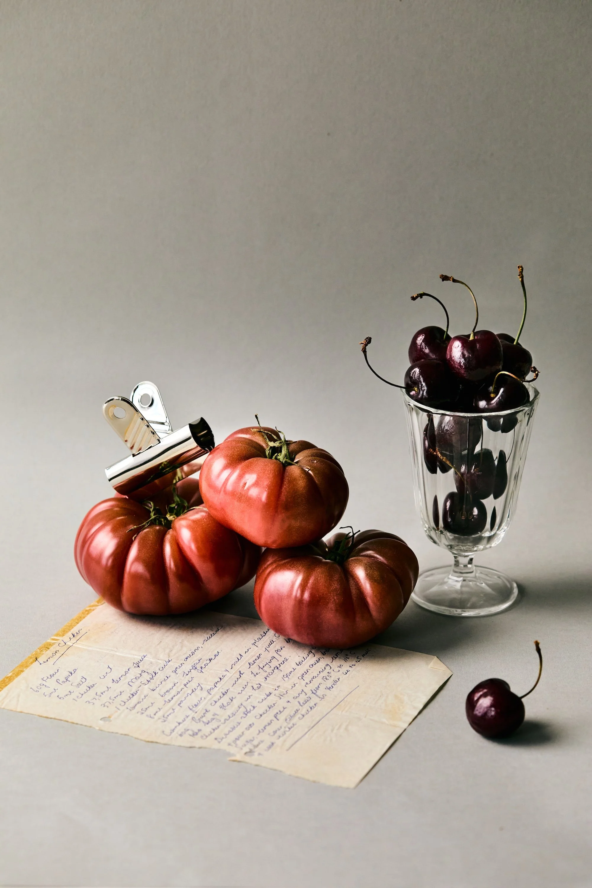 Tomatoes with a paper clip, a wine glass of cherries, a single cherry on the surface, and a handwritten letter on a flat surface.