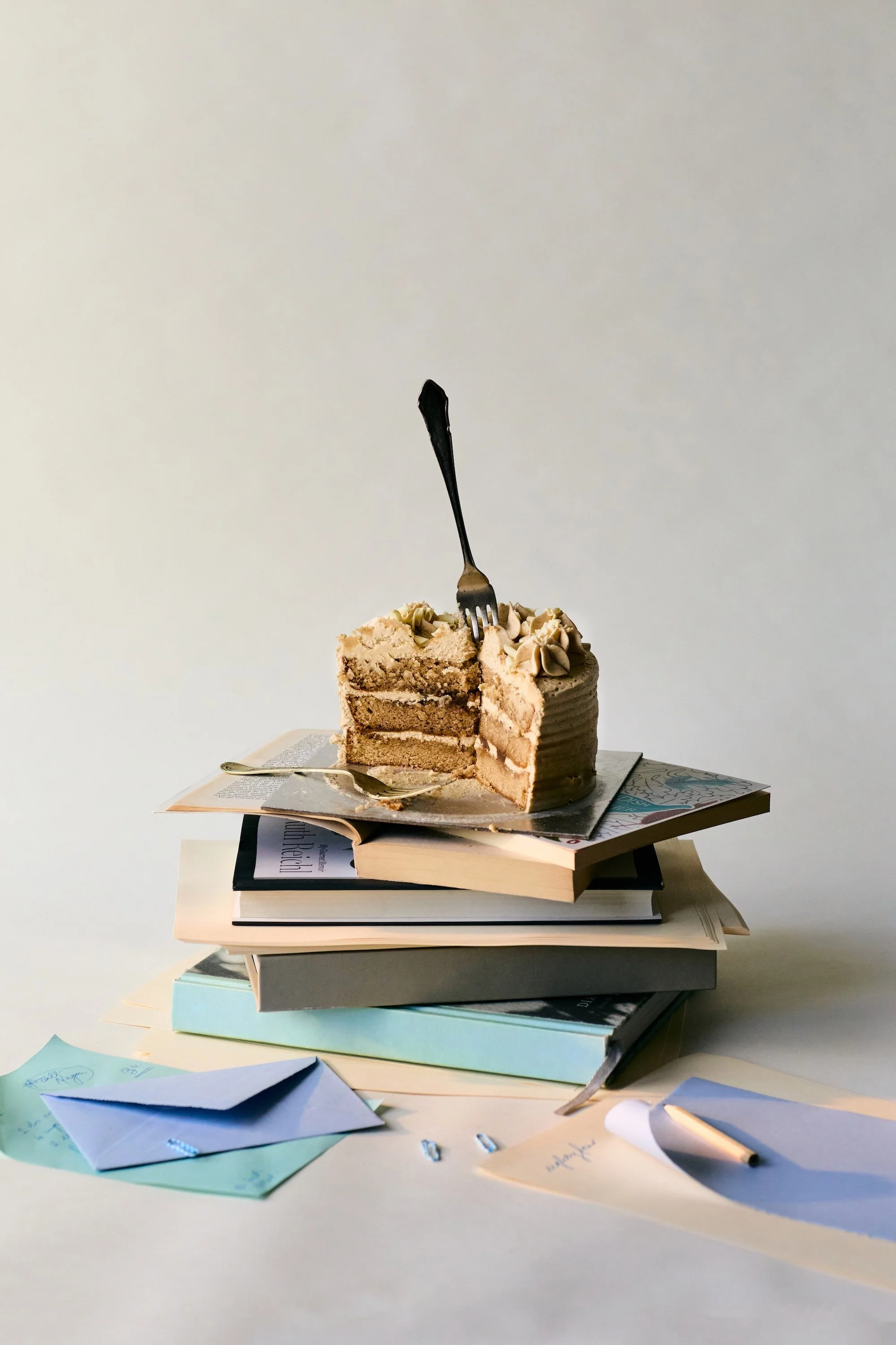 A stack of books with a partially eaten slice of layered cake on top, a fork inserted into the cake, and scattered papers and envelopes around.