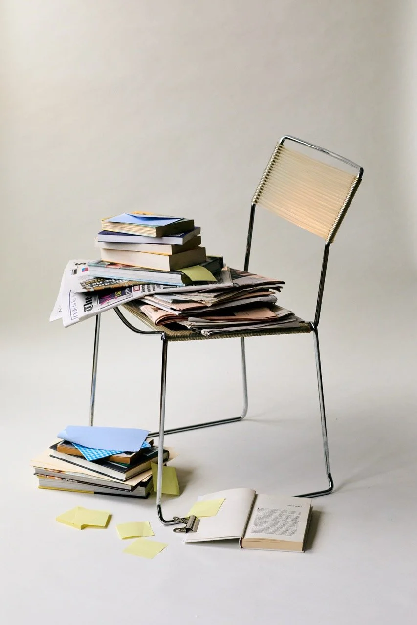 A metal chair with a beige woven seat and backrest, cluttered with stacks of papers, notebooks, and folders, with some papers and sticky notes on the floor and an open book at the base.