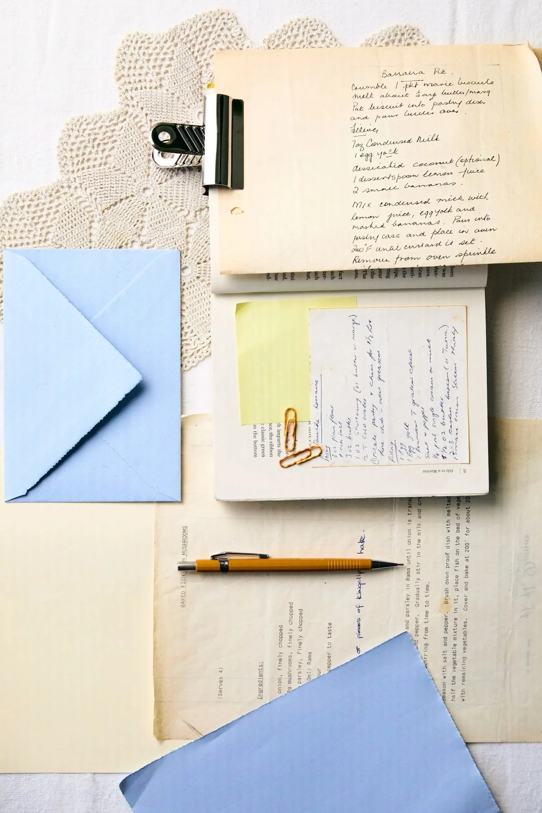 A flat lay of a workspace includes a notebook with handwritten recipes, a yellow pen, a blue envelope, a note with handwritten text, photo paper clips, and a lace doily on a white surface.