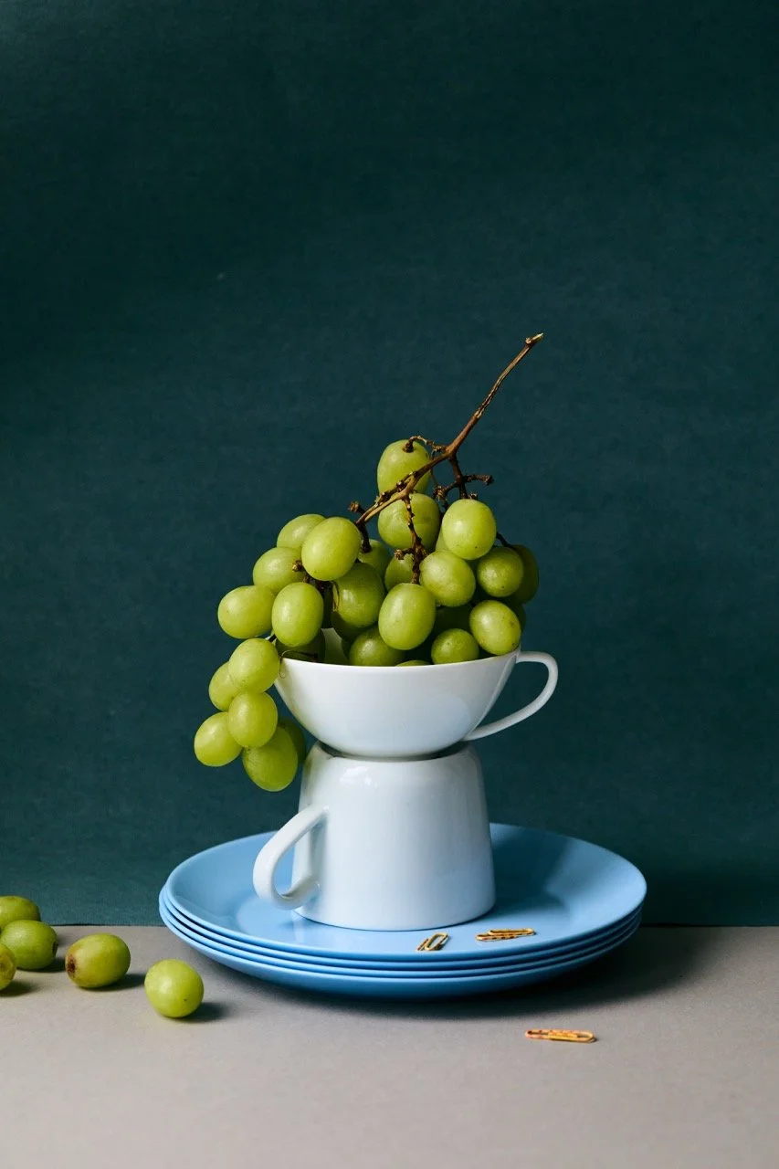 Still life of green grapes in a white mug on a stack of light blue plates, with a few grapes and paper clips on the surface, and a dark teal background.