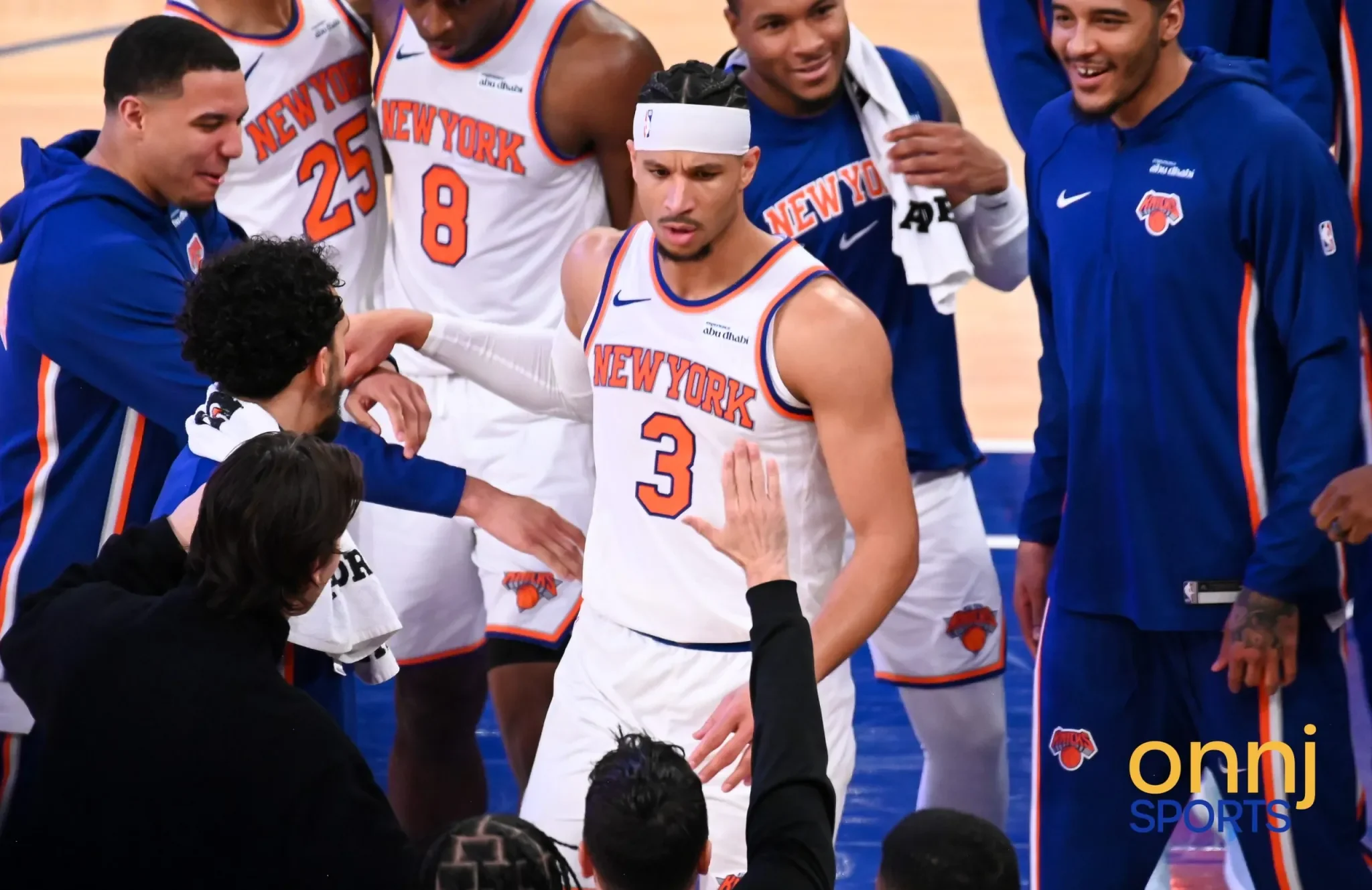 A group of New York Knicks basketball players and staff gathered on the court, some wearing team jerseys and others in team jackets, with a young player wearing a headband in the center, appearing focused or serious.