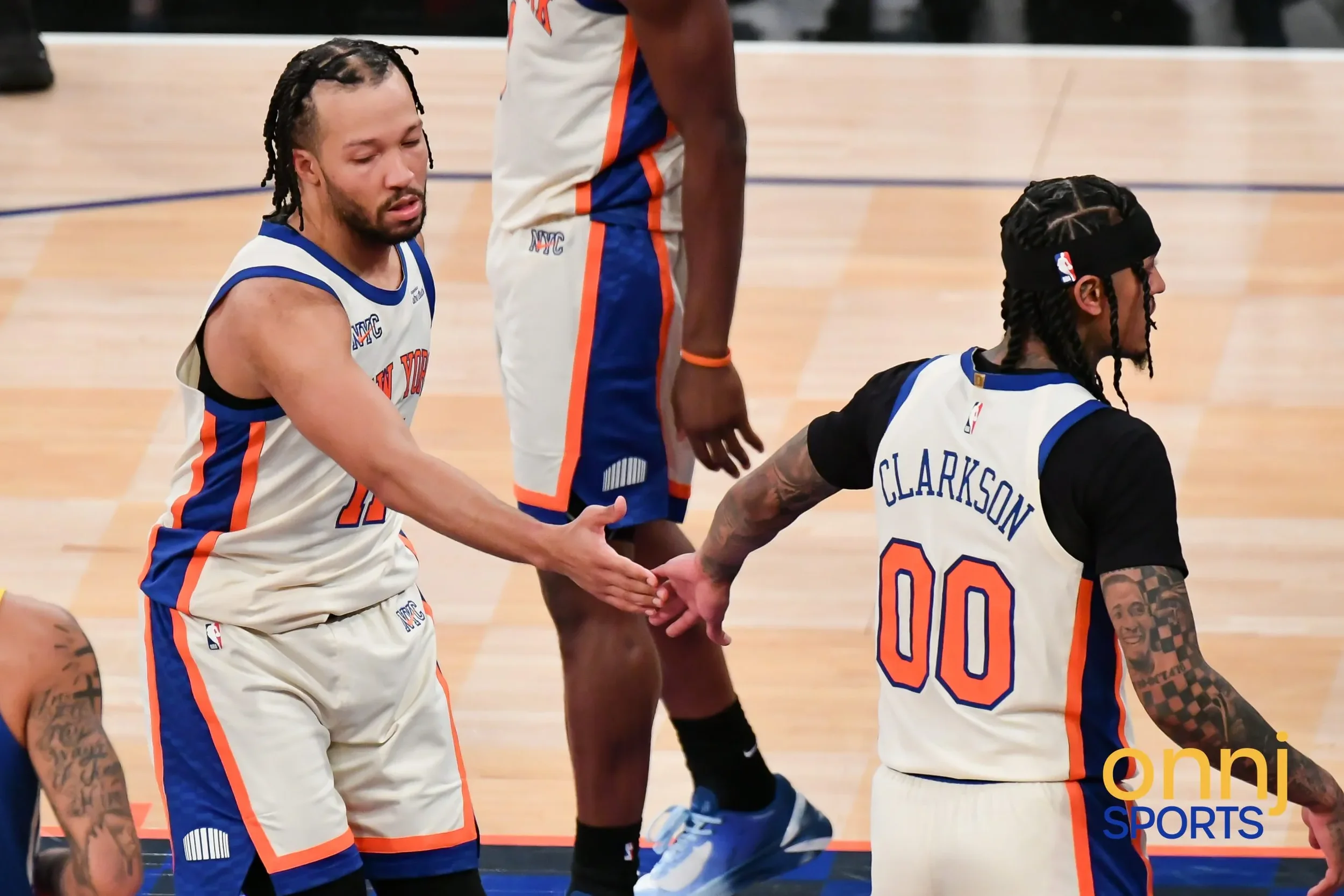 Jalen Brunson and Jordan Clarkson from the New York Knicks shake hands on the court, wearing white jerseys with blue and orange accents, during a game.