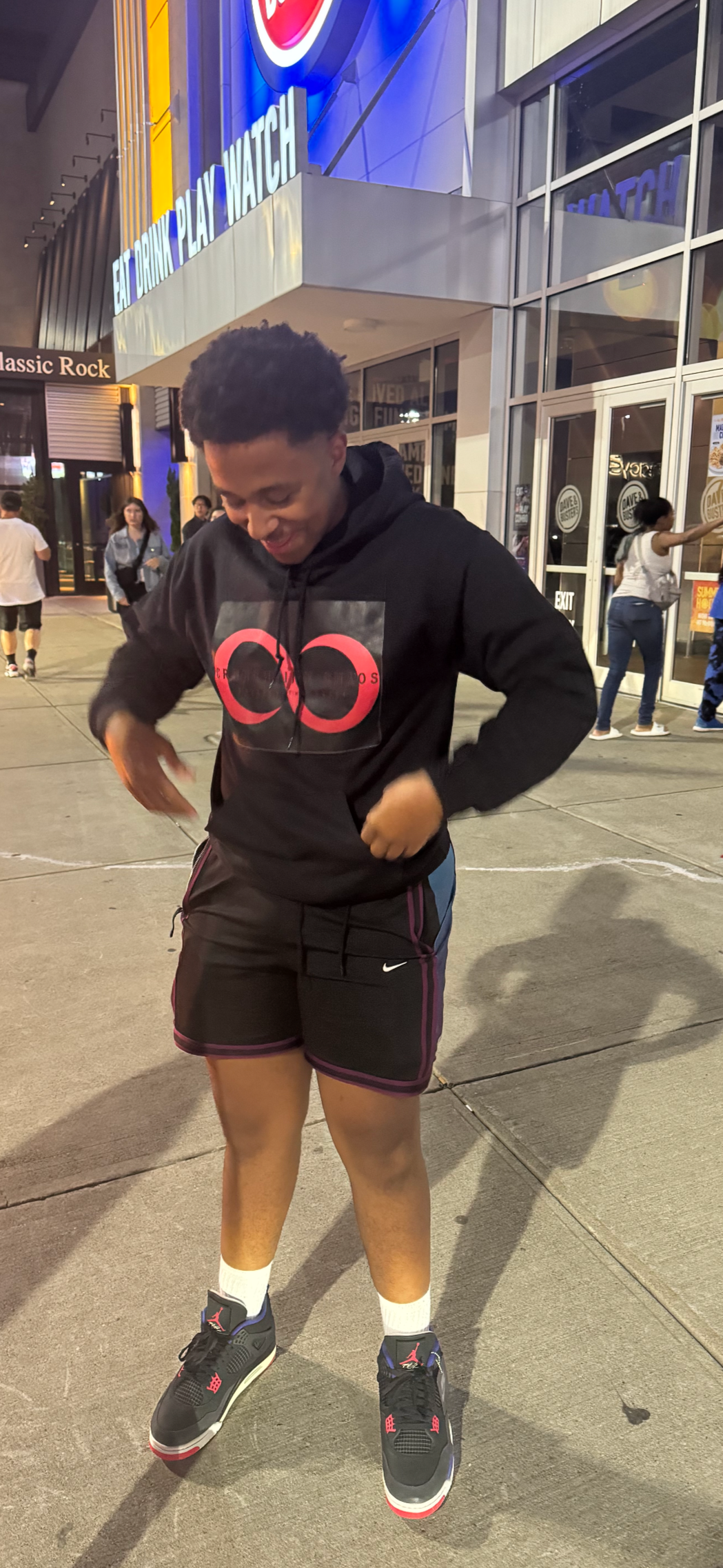 Young man standing outside a retail store at night, wearing a black hoodie, black shorts, and black sneakers, smiling and looking downward.
