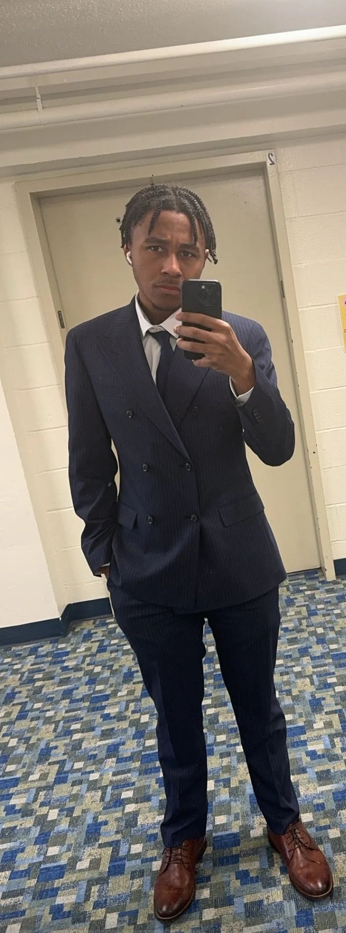 Young man in a navy blue double-breasted suit taking a mirror selfie in a hallway with beige walls and patterned carpet.