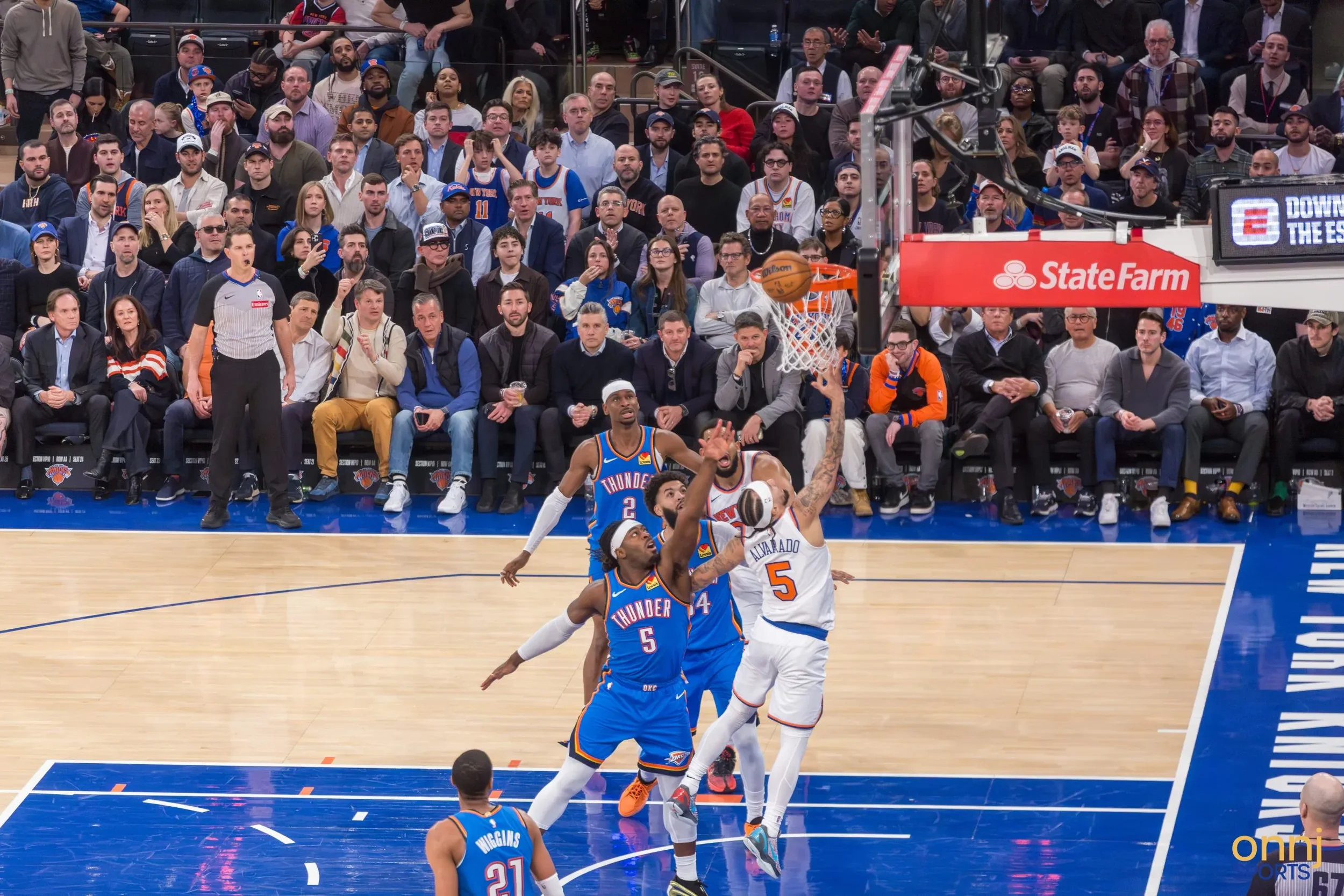 A basketball game with players from the Oklahoma City Thunder competing against the New York Knicks. A player from the Knicks wearing jersey number 5 is attempting a shot while being defended by a player from the Thunder. The court is filled with spectators watching the play.